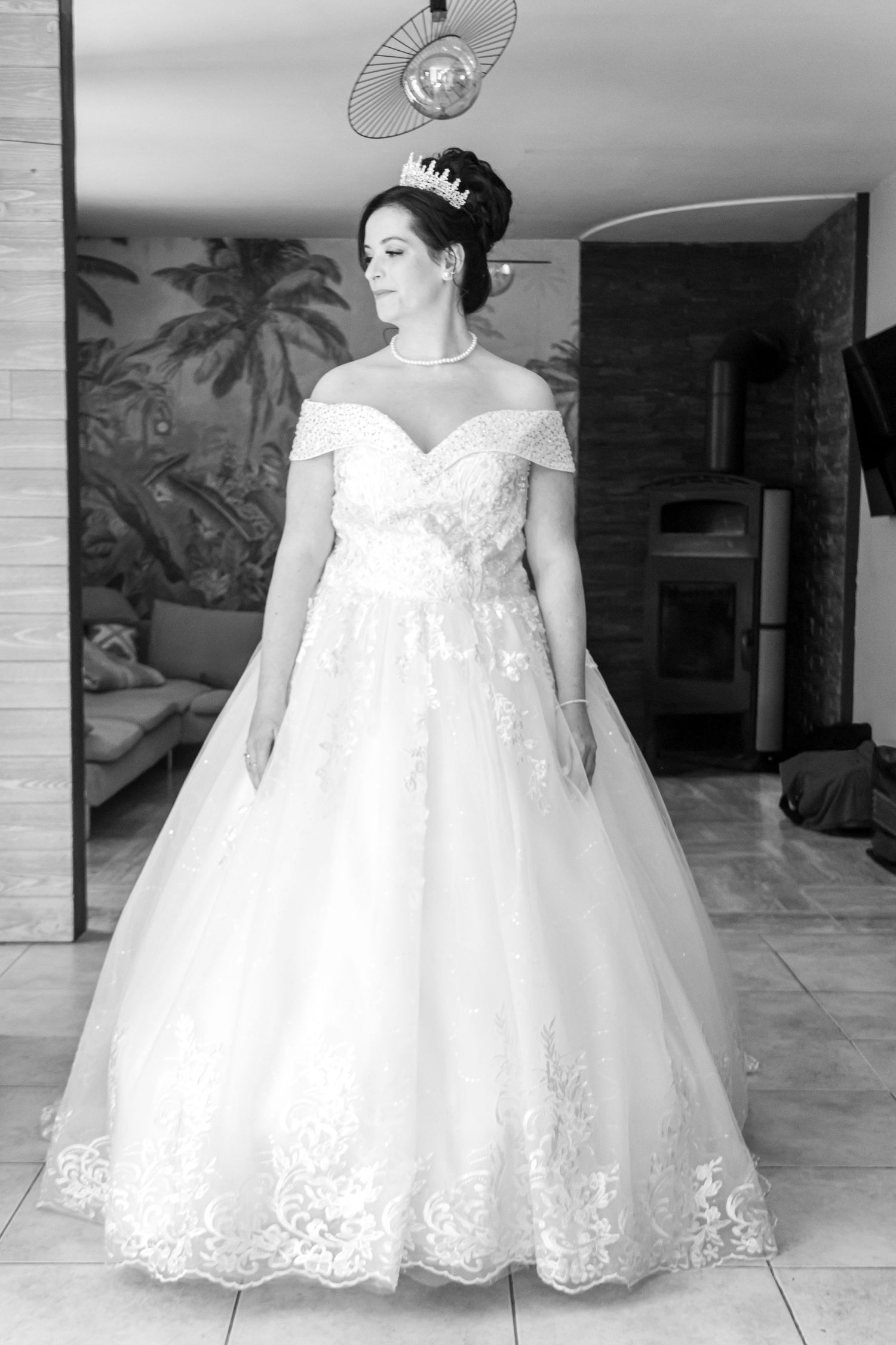 Bride in elegant off-shoulder ball gown wedding dress with lace details and tiara, posing indoors in black and white photo.