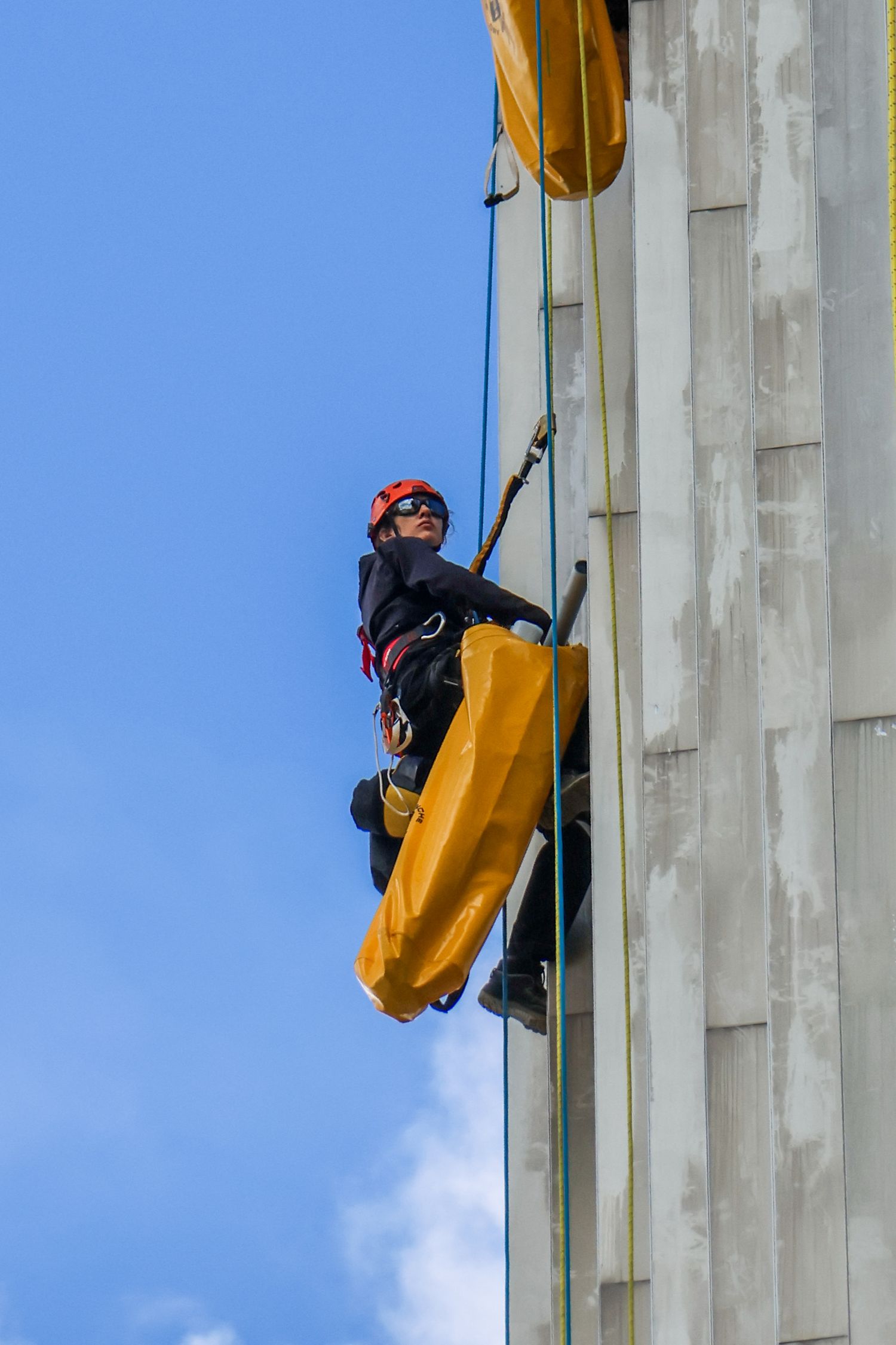 A worker in red helmet and black gear rappels down a concrete building wall against a blue sky.