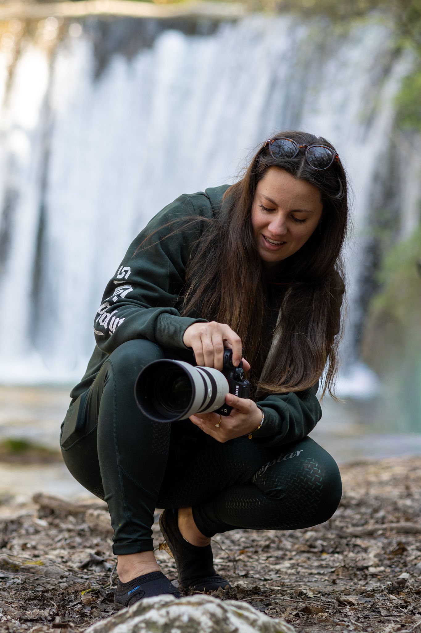 A woman crouches near a waterfall, smiling while adjusting a professional telephoto camera lens outdoors.