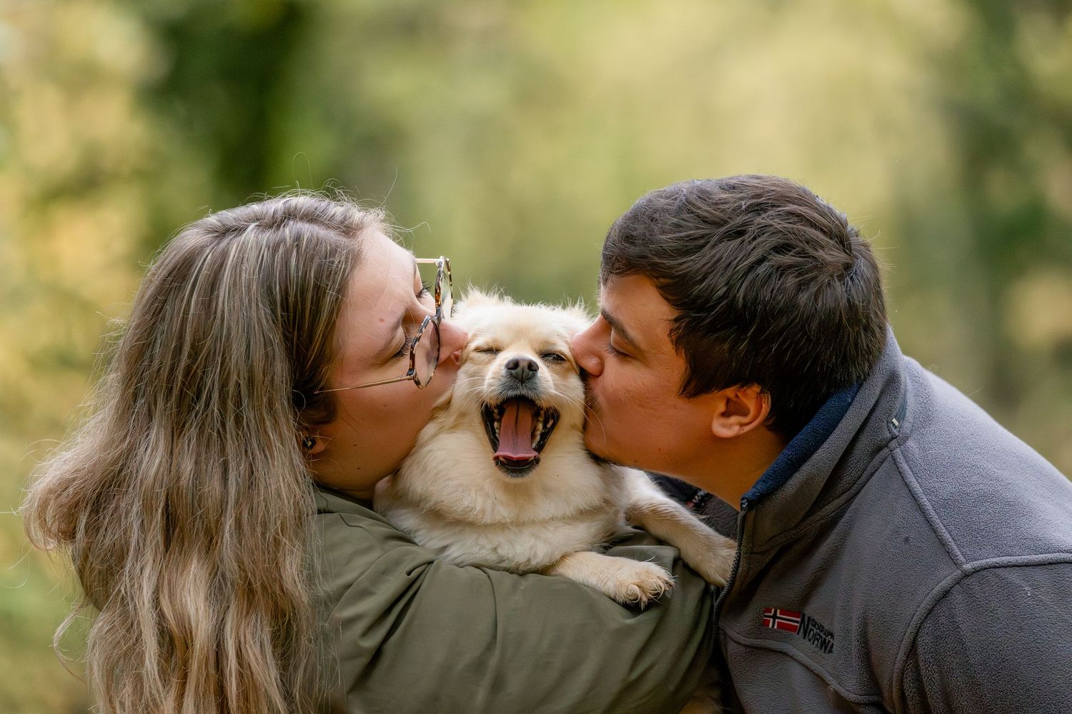 A couple kisses their happy Pomeranian dog outdoors in a lush green forest setting.