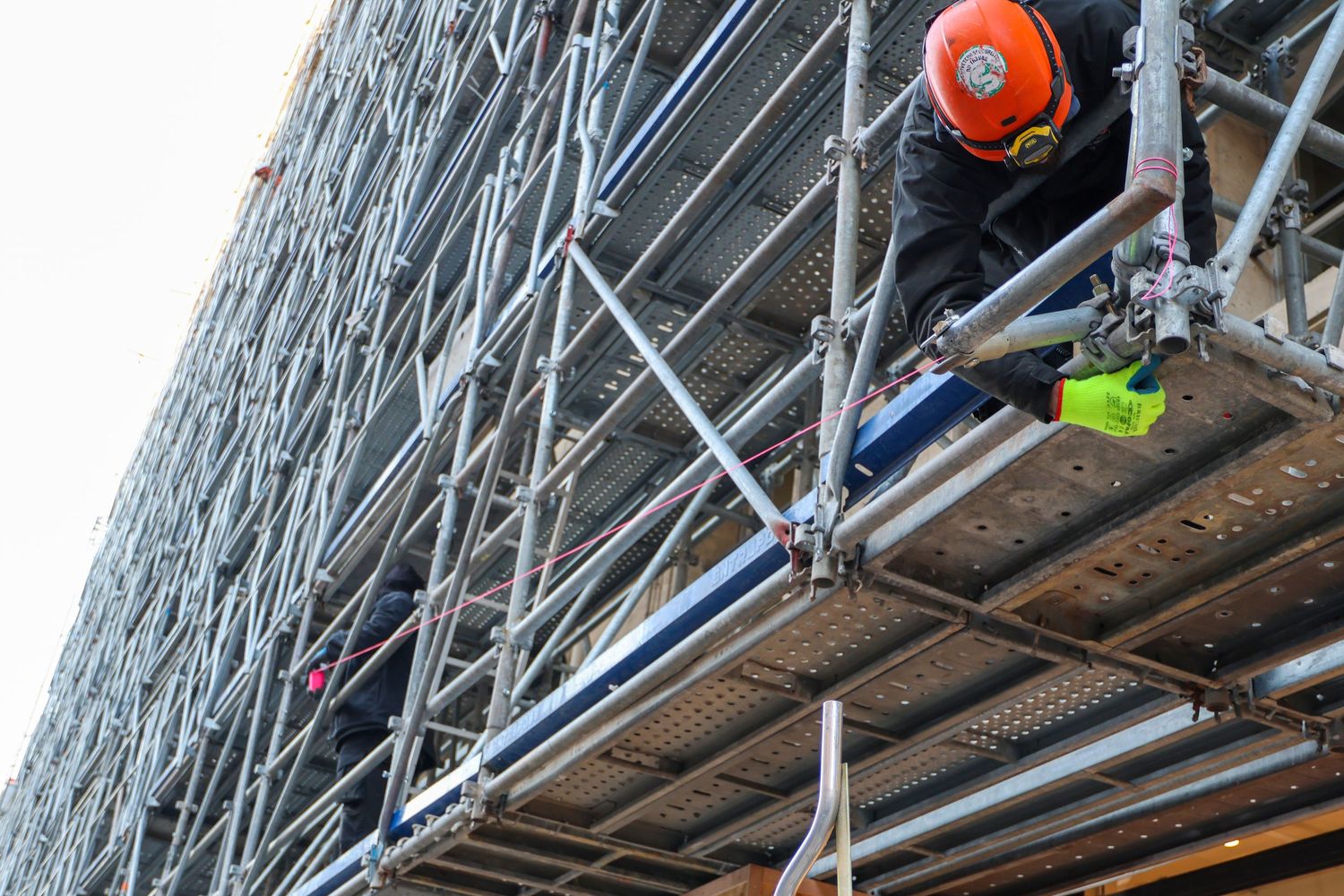 Construction worker in orange helmet on scaffolding inspecting a building facade at height.