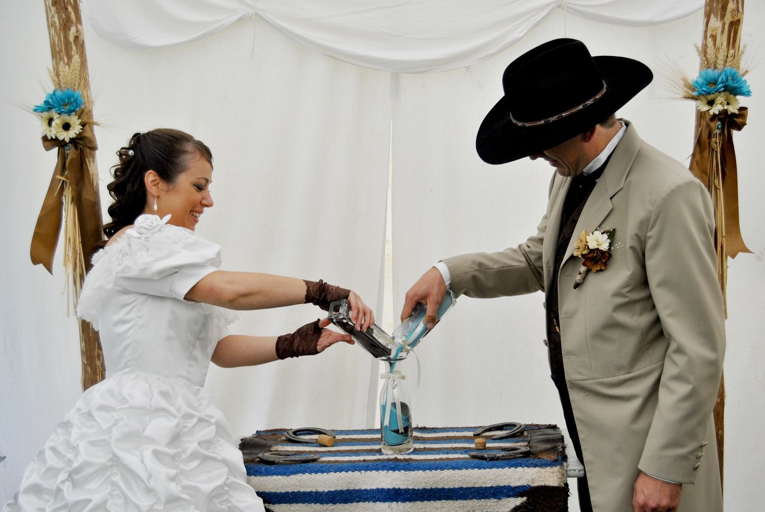 Bride and groom in cowboy-themed attire pour sand into a vase during a unity ceremony at an outdoor wedding.