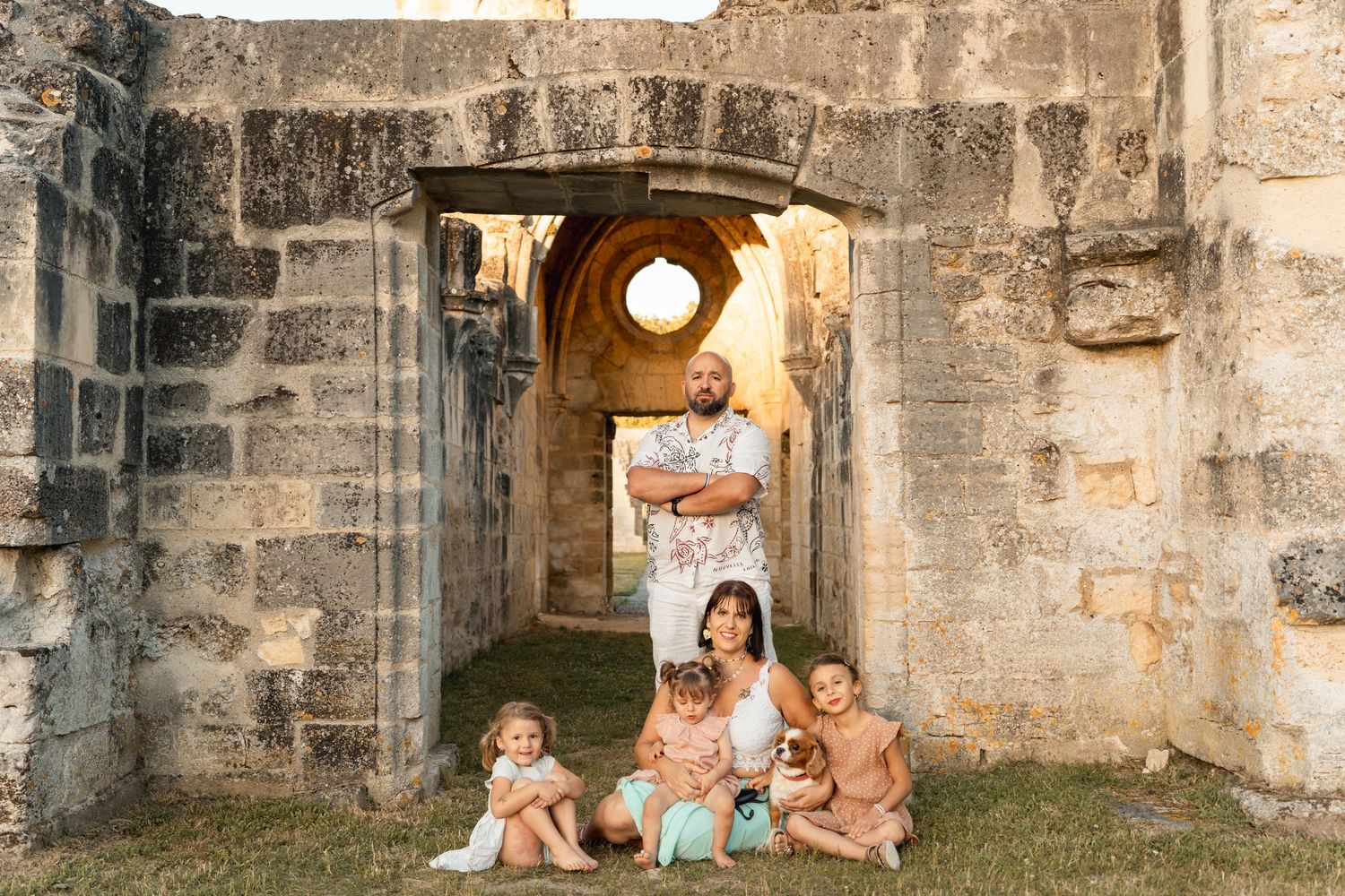 Family portrait with parents and three young children sitting together in front of a stone archway ruin.
