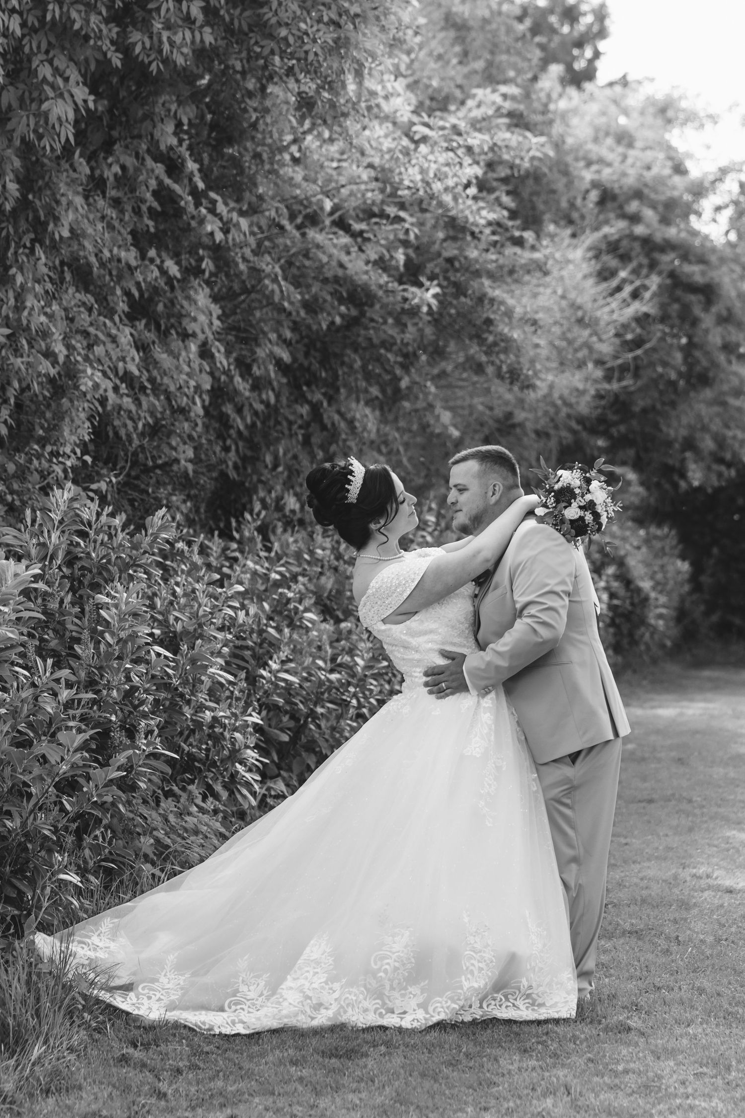 Black and white wedding photo of bride and groom embracing in a lush garden, bride in elegant lace gown holding bouquet.