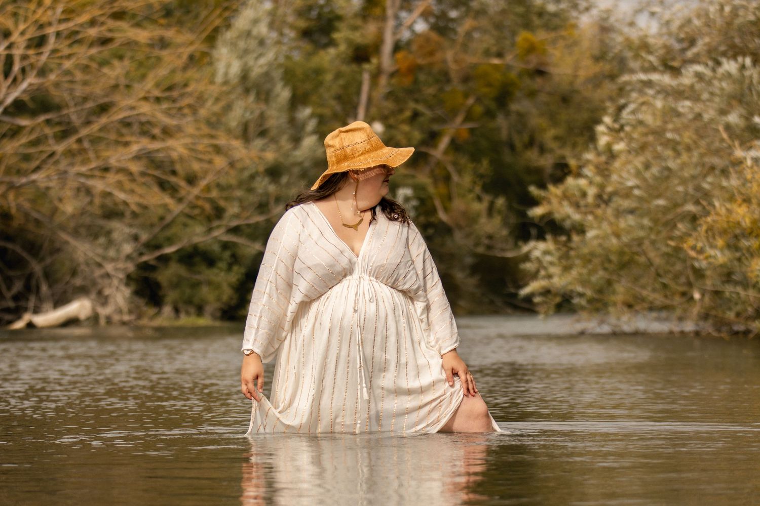 Pregnant woman in white dress and straw hat standing in a serene river surrounded by autumn trees.
