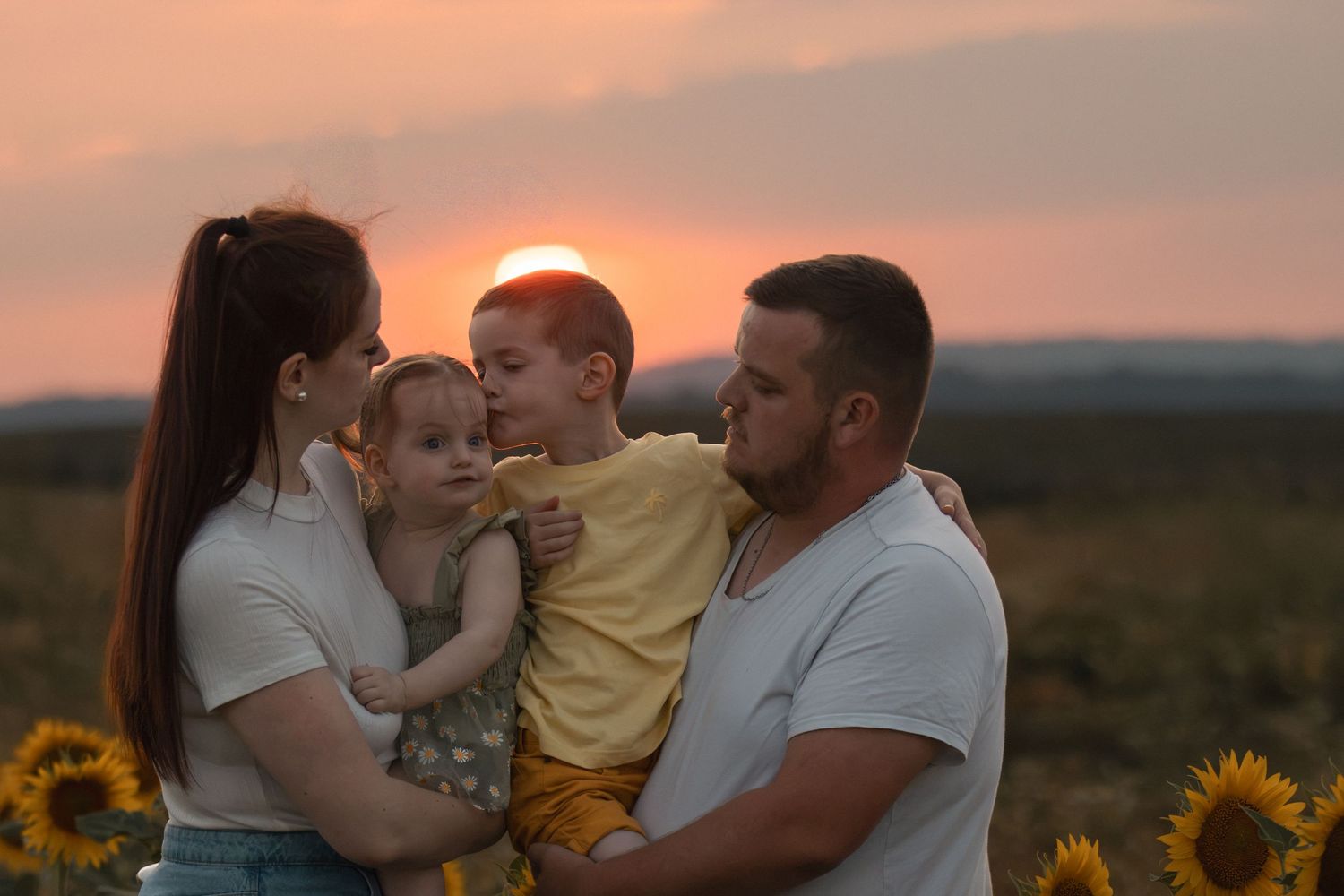 Family of four enjoying a golden sunset in a sunflower field, parents holding young children outdoors.