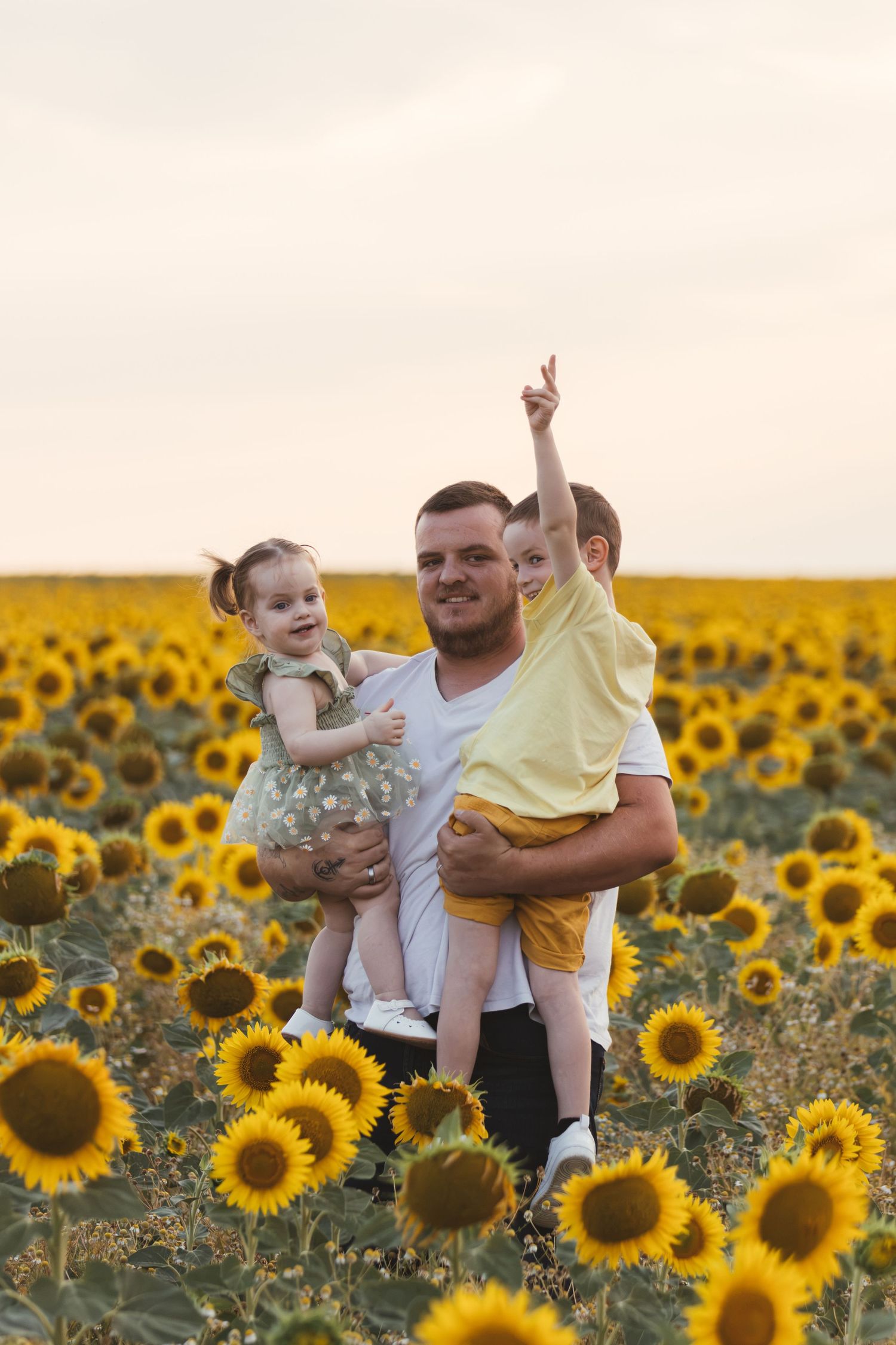 Father holding two young children in vibrant sunflower field at sunset.
