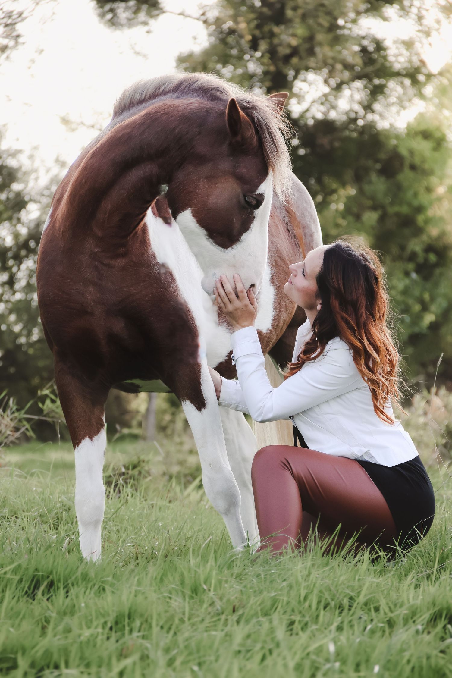 Woman in white jacket kneeling beside a brown and white paint horse on green grass.