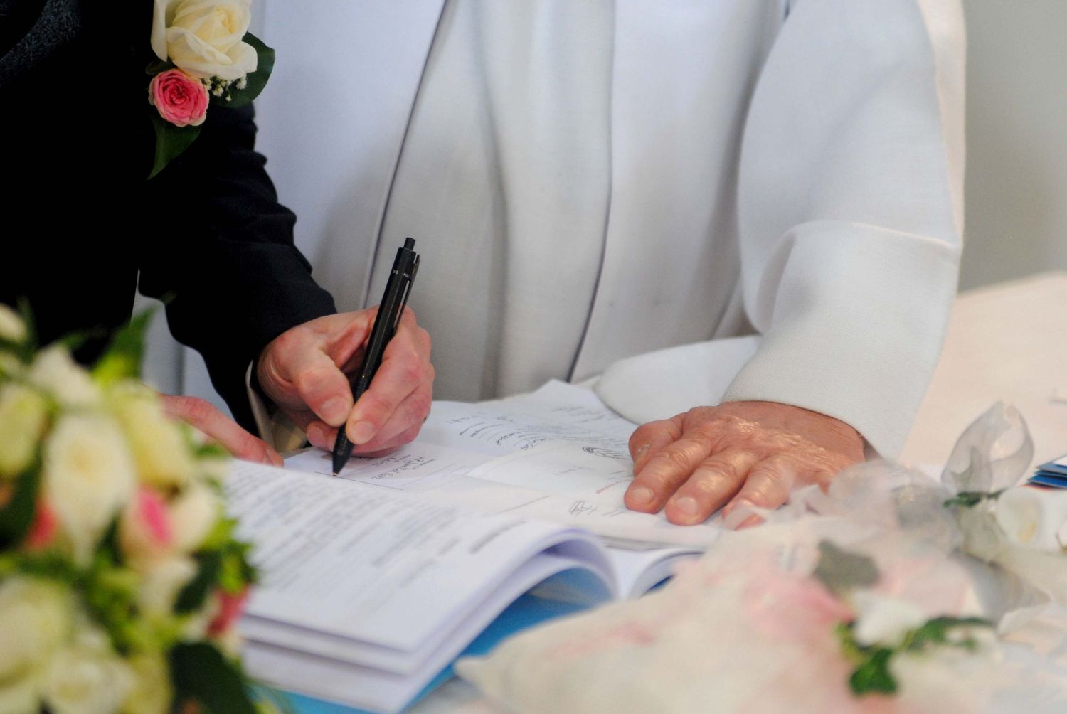 Bride signing wedding register document with pen at ceremony table decorated with pink and white flowers.
