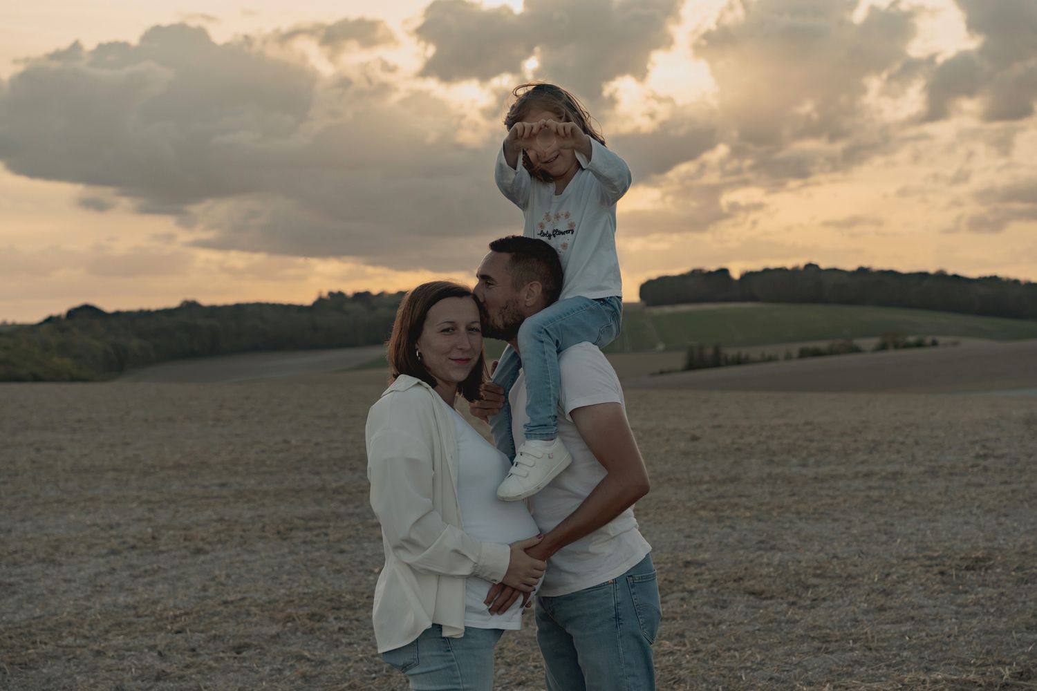 Family of three posing at sunset in an open field, parents smiling as child sits on fathers shoulders.