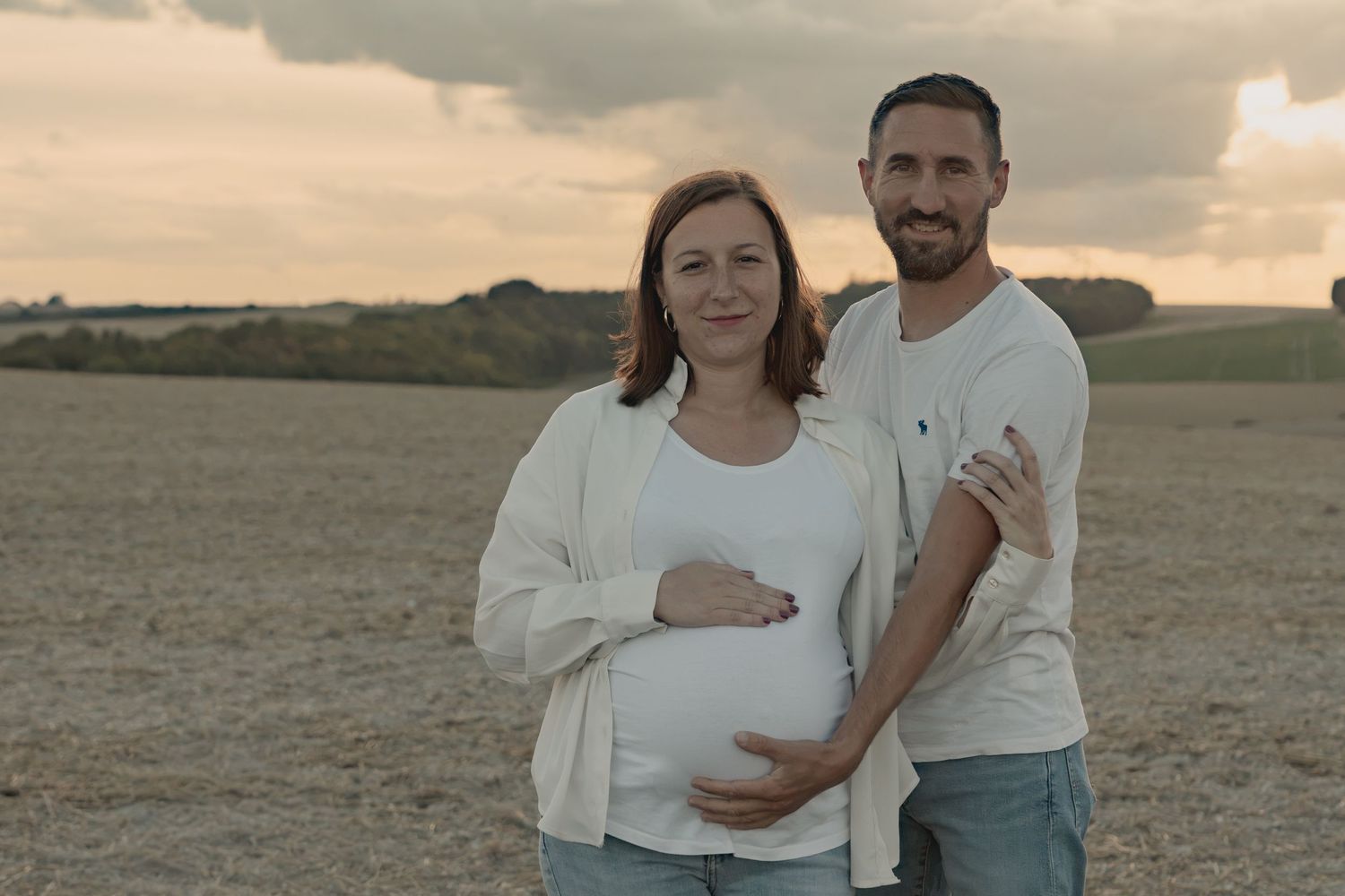 Pregnant couple posing together in an open field at sunset, woman cradling baby bump, man embracing her.