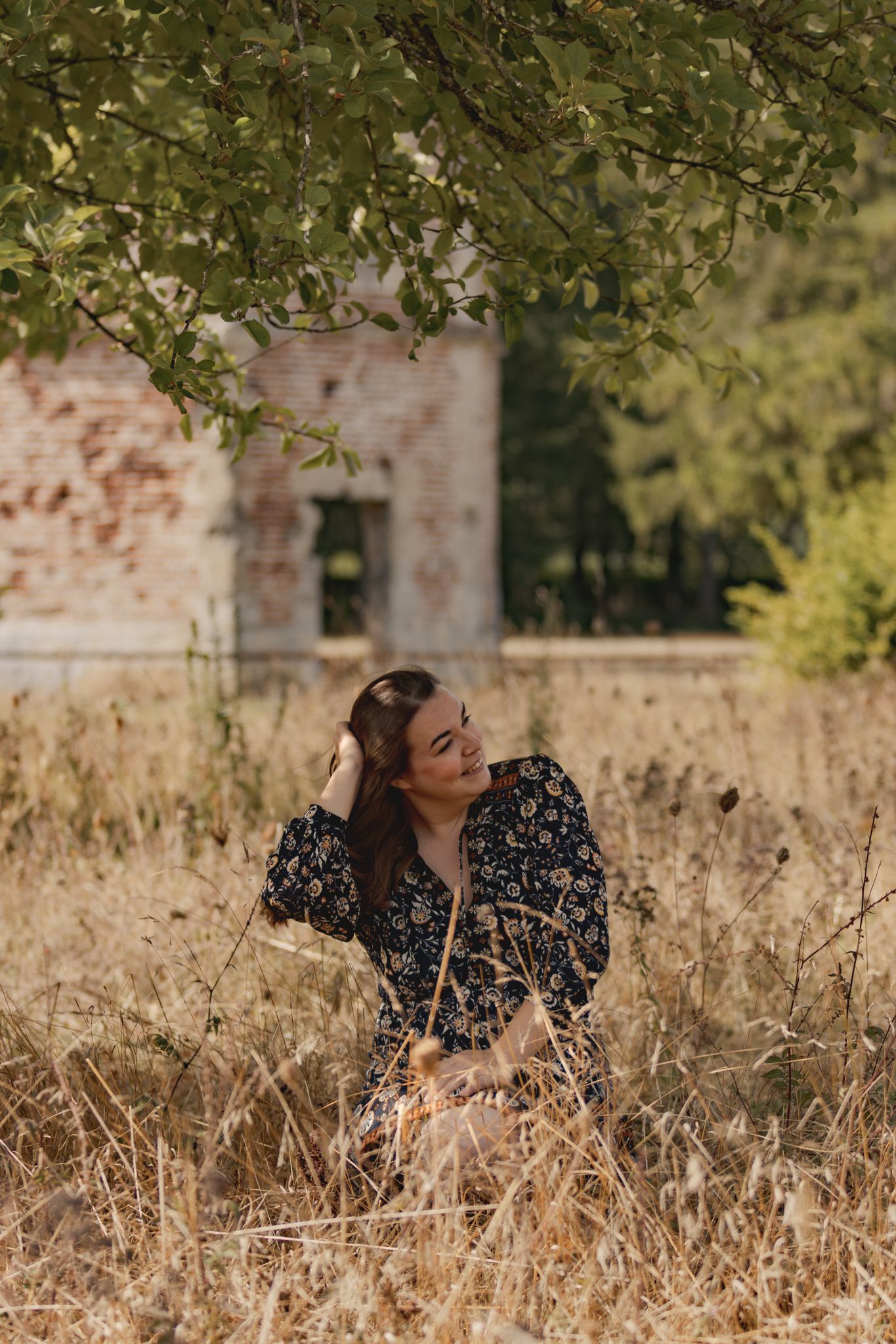 Woman in floral dress sitting in golden field beneath tree branches near rustic brick building.