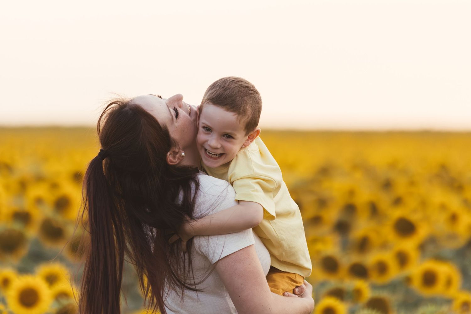 A mother kissing her laughing son in a vibrant sunflower field at golden hour.