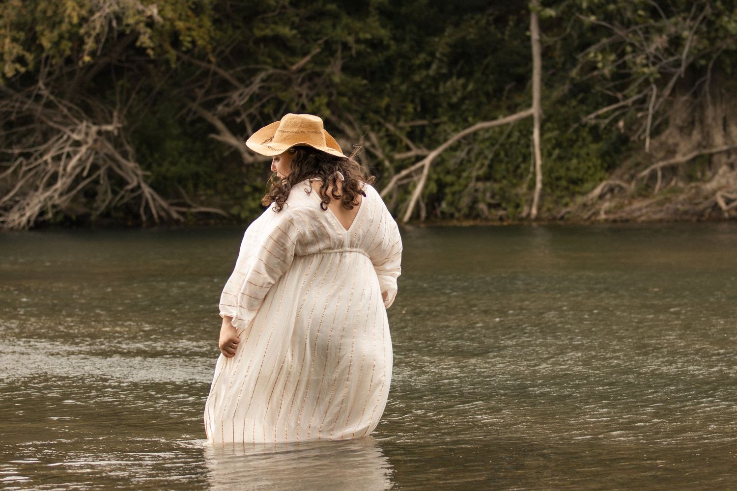 Pregnant woman in white dress and straw hat wading in a river, back facing camera, surrounded by trees.
