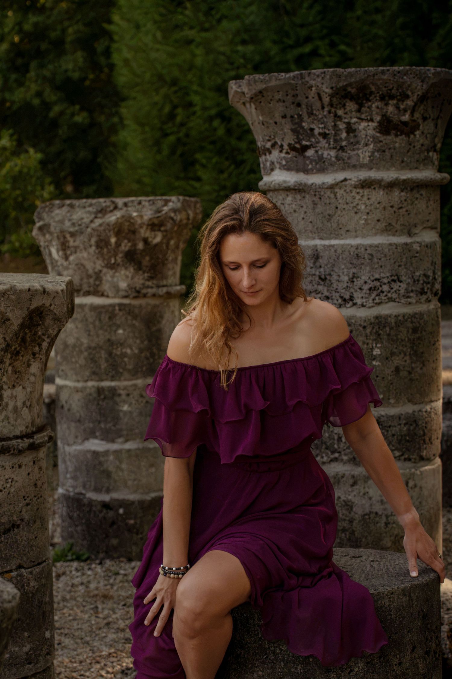 Woman in purple off-shoulder ruffled dress sitting on stone steps surrounded by ancient columns and green trees.