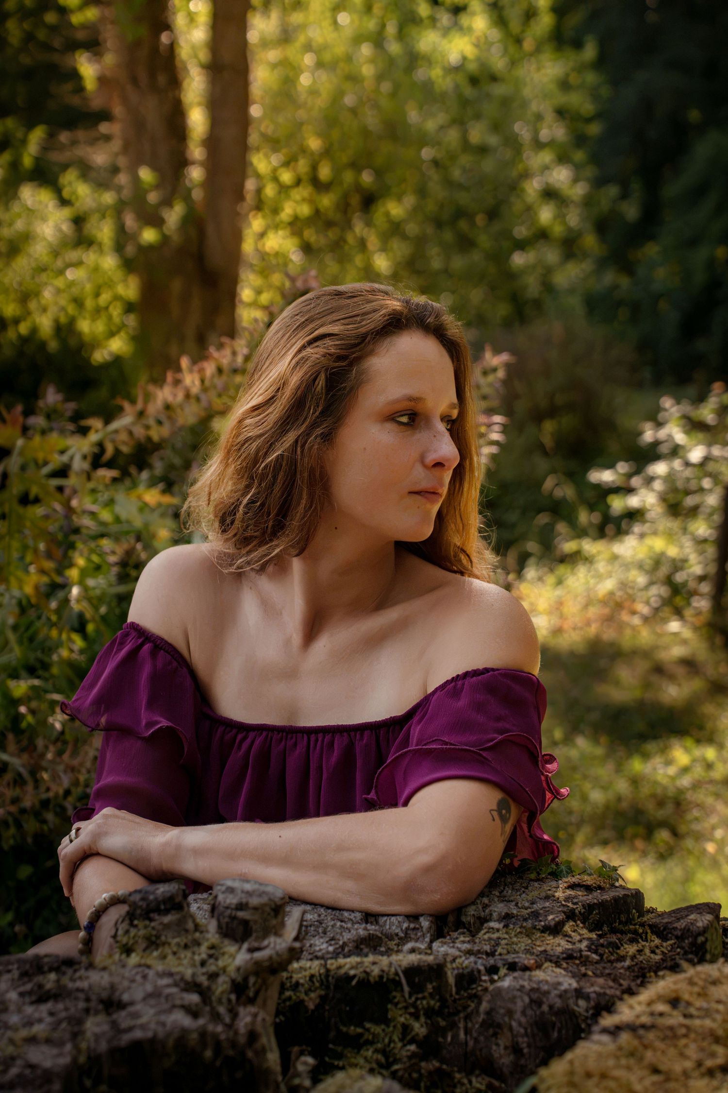Woman in purple off-shoulder dress leaning on mossy rocks in a sunlit forest setting.