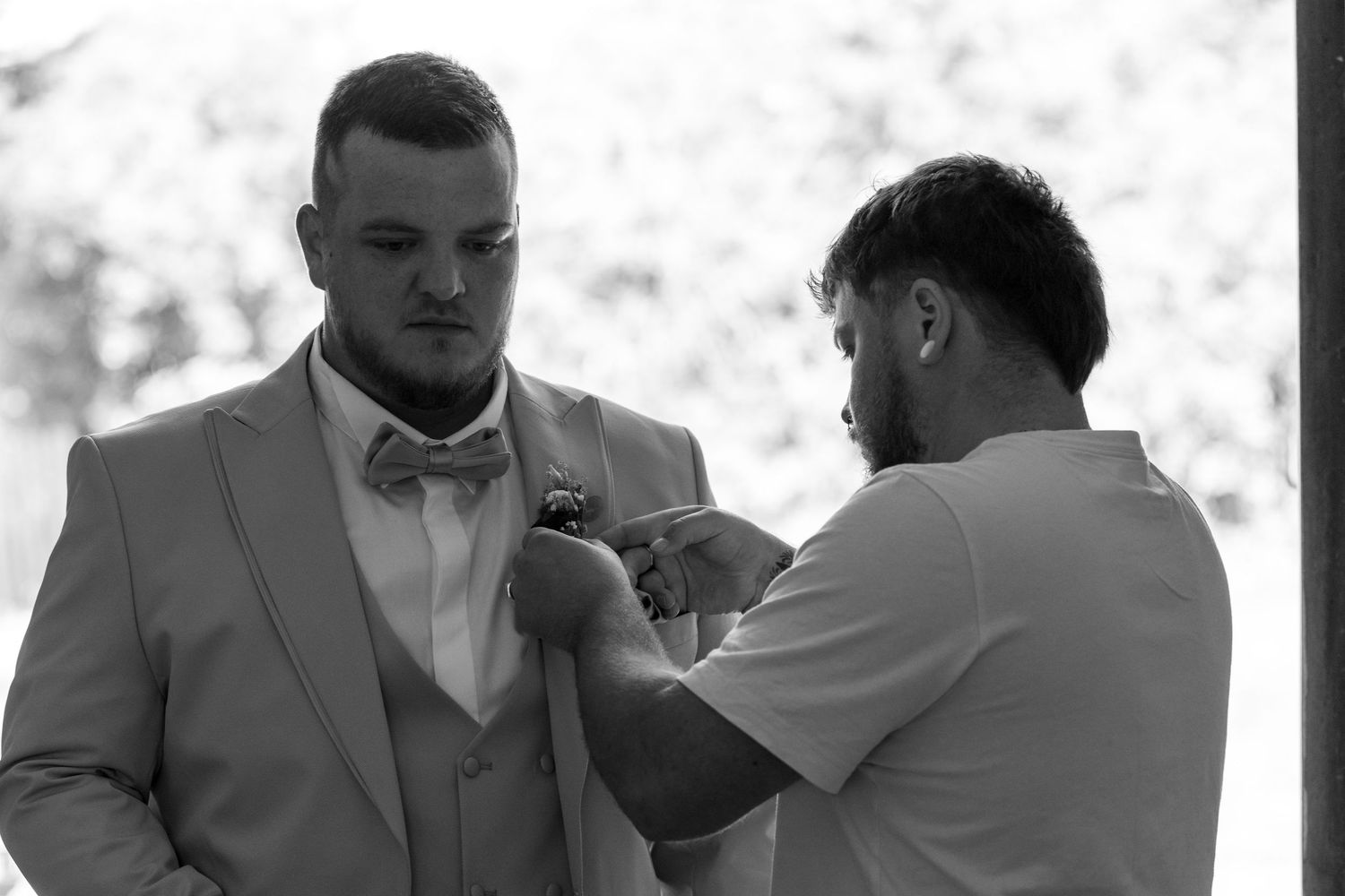 Black and white photo of a groom in a suit and bow tie having his boutonniere pinned on by a groomsman.
