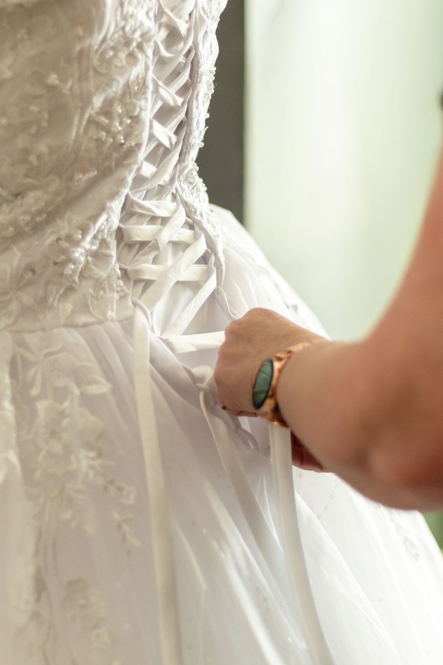 A hand with a gold bracelet lacing up the back of a white wedding dress with intricate lace detailing.