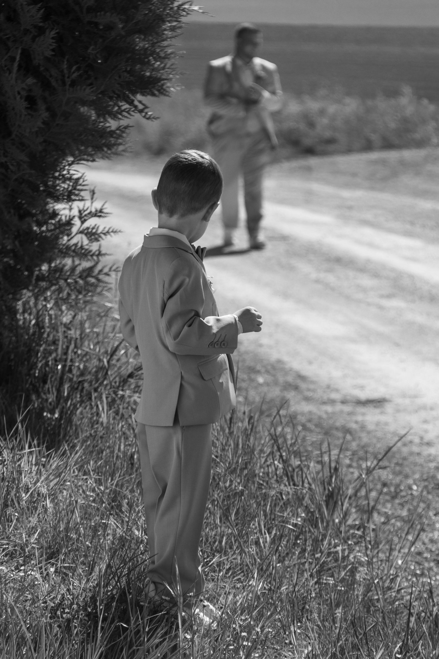 Black and white photo of a young boy in a suit standing on a rural dirt road with a man in the background.