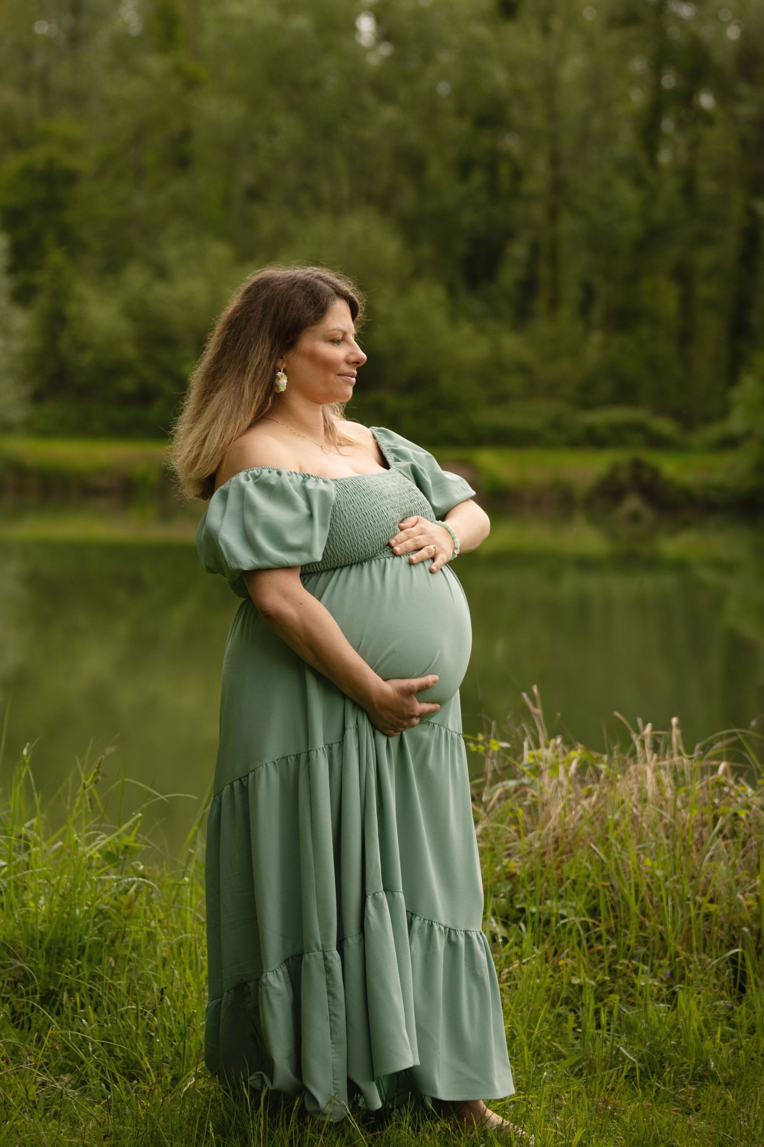 Pregnant woman in sage green off-shoulder dress cradling baby bump outdoors by a serene pond surrounded by lush greenery.
