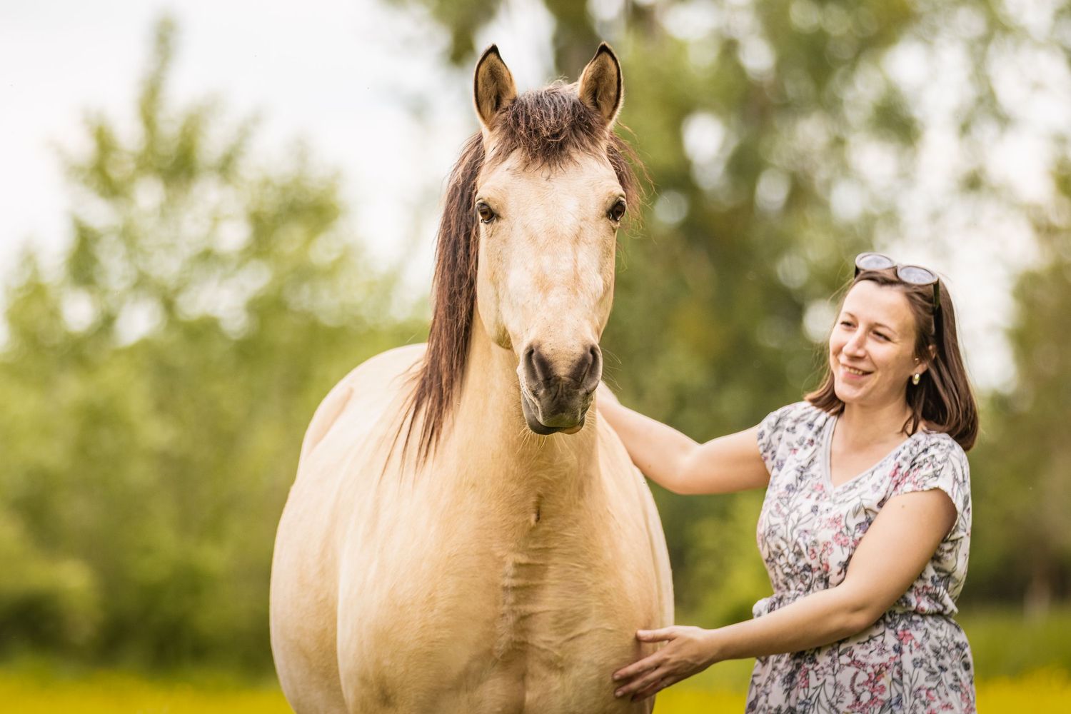 Smiling woman in floral dress petting a golden palomino horse outdoors in a green field.