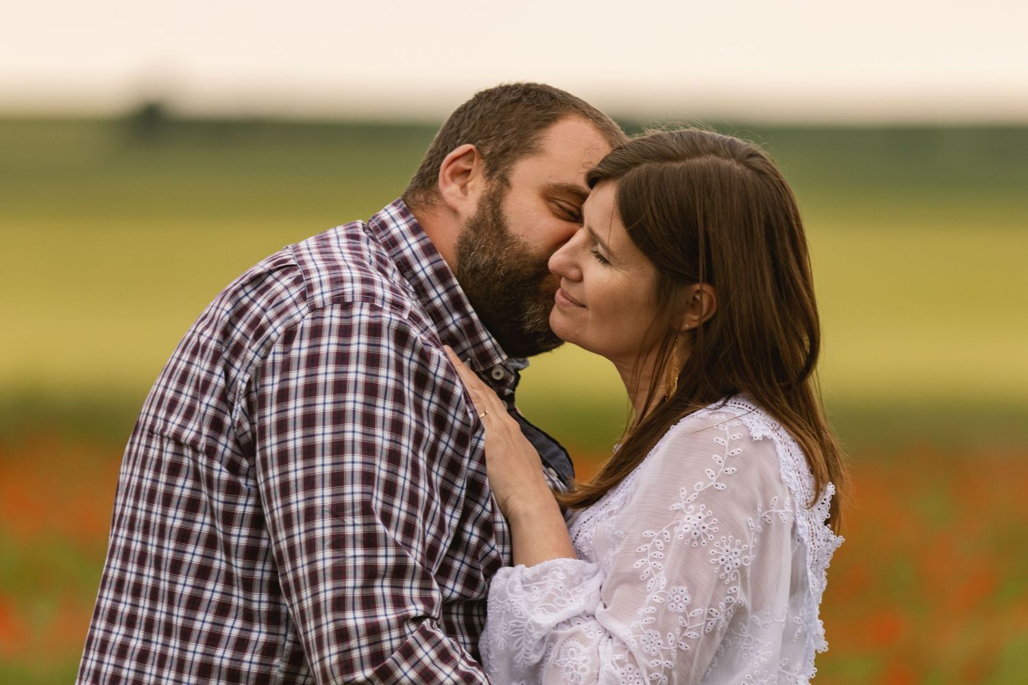 Romantic couple sharing a tender moment in a colorful flower field at sunset, man kissing woman's forehead.