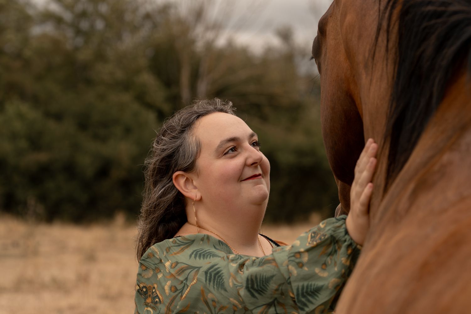 Woman smiling while gently touching a brown horse outdoors with trees in the background.