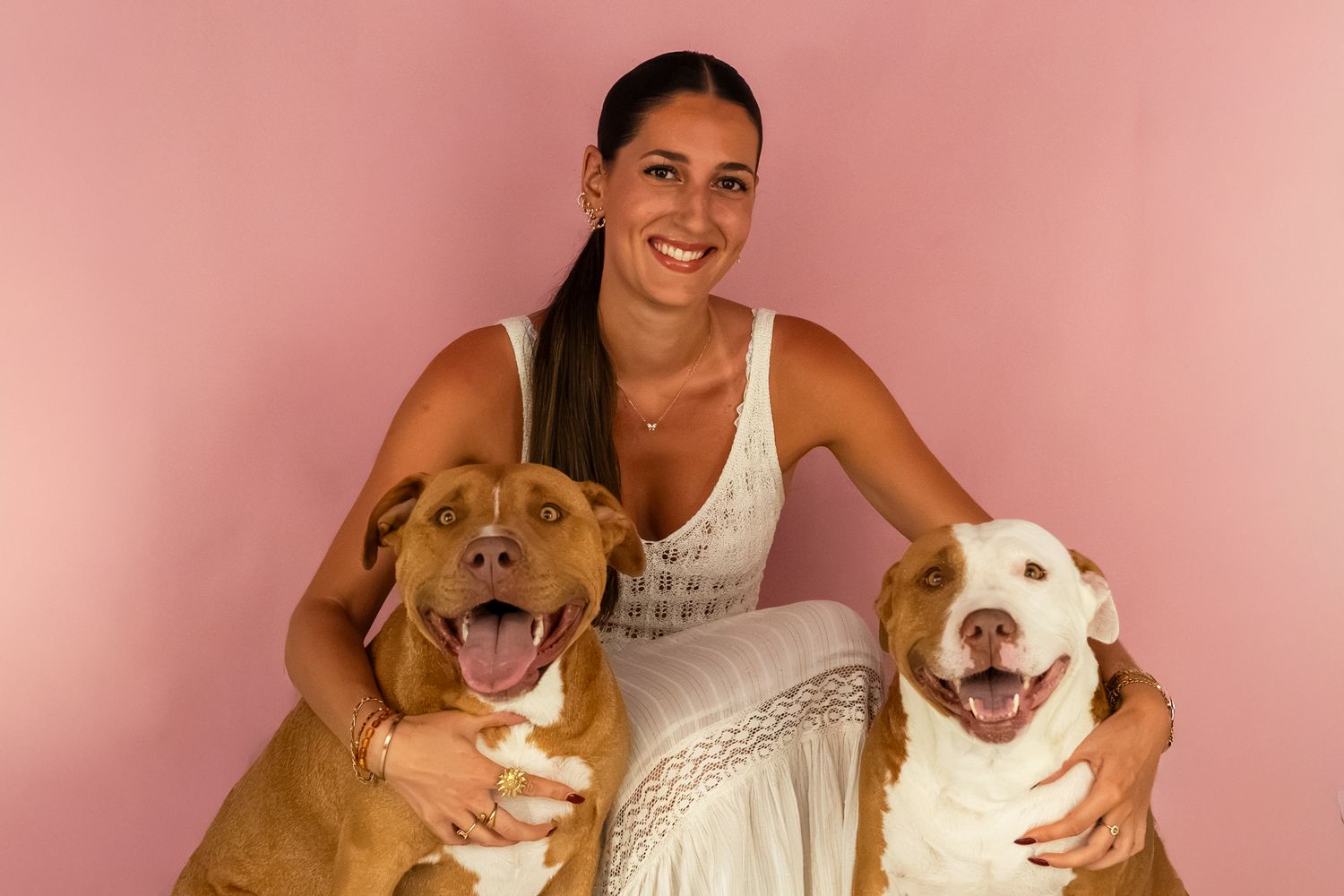 Woman in white dress smiling with two pit bull dogs against a pink background.