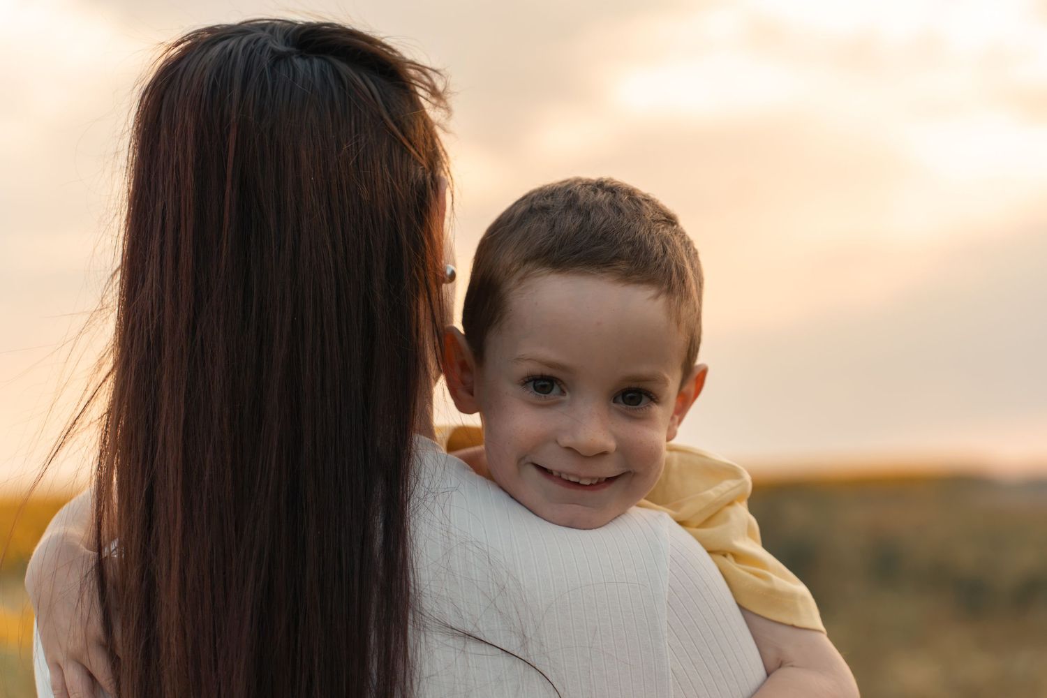 A mother holds her smiling toddler boy outdoors at sunset, the child looking over her shoulder with a joyful expression.