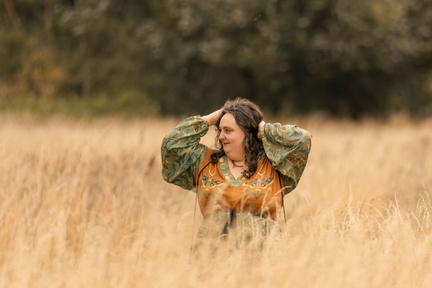 Woman in colorful floral blouse standing in golden grass field with hands in hair, looking sideways outdoors.