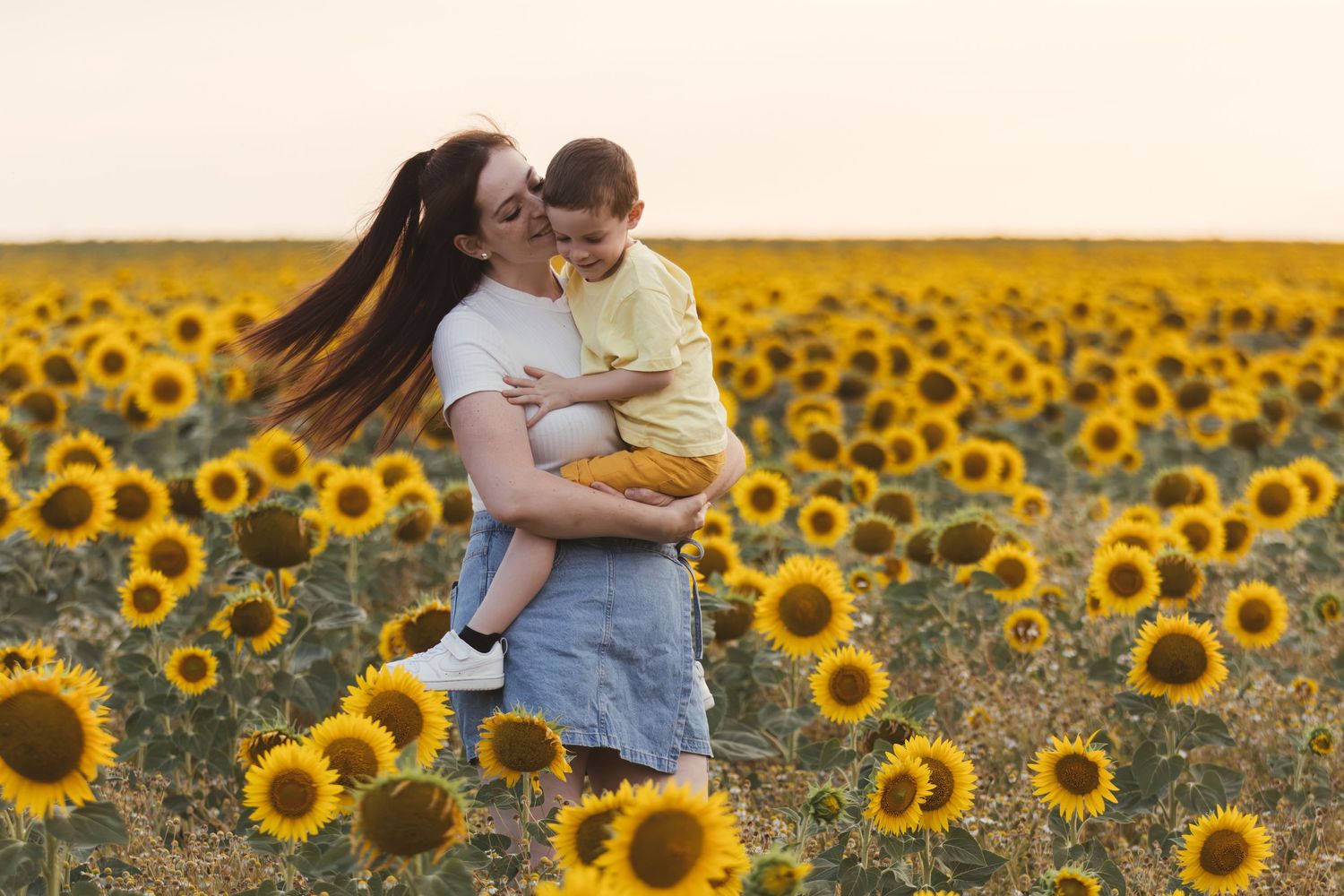 A mother holds and kisses her toddler in a vibrant sunflower field at sunset.