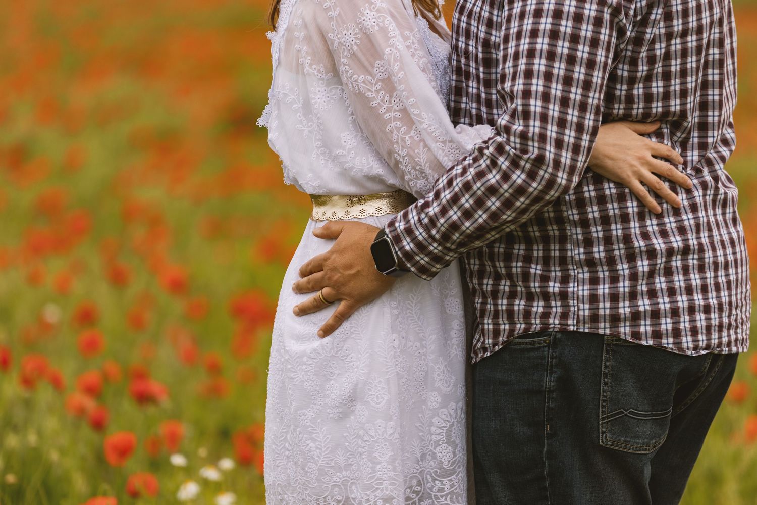 Couple embracing in a vibrant red poppy field, woman in white lace dress and man in plaid shirt.