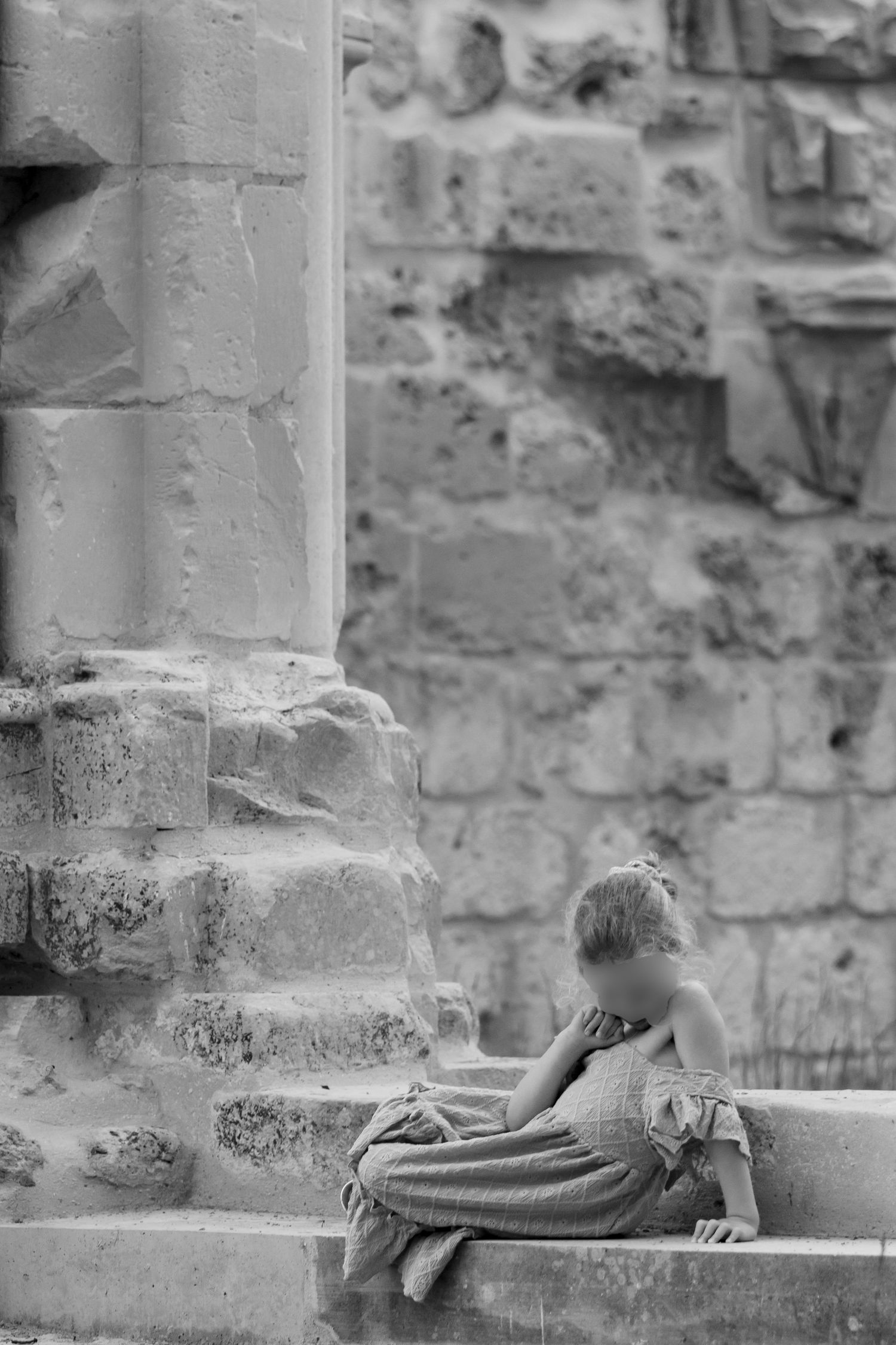 Black and white photo of a young girl sitting alone on stone steps beside an ancient column.