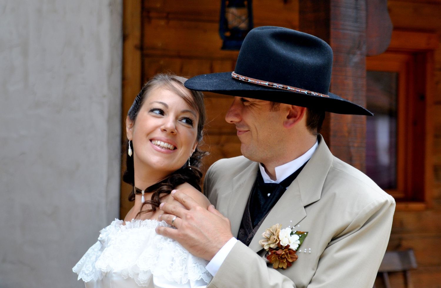 Smiling western bride in white gown embraced by groom in tan suit and black cowboy hat outdoors.