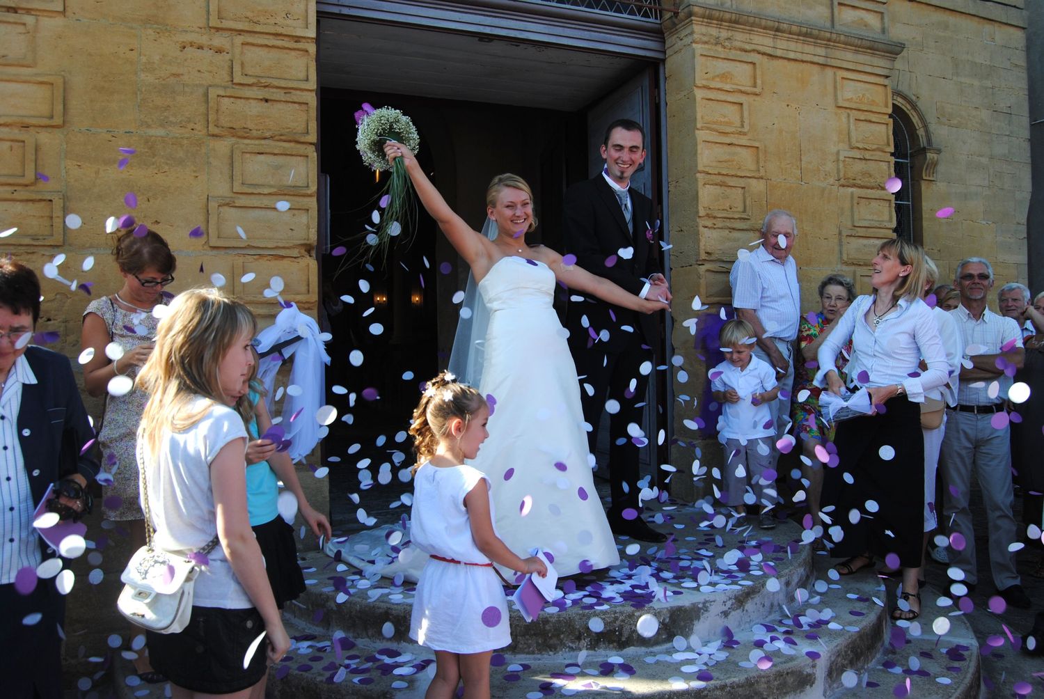 Bride and groom exit church doors as guests toss colorful confetti during a sunny outdoor wedding celebration.