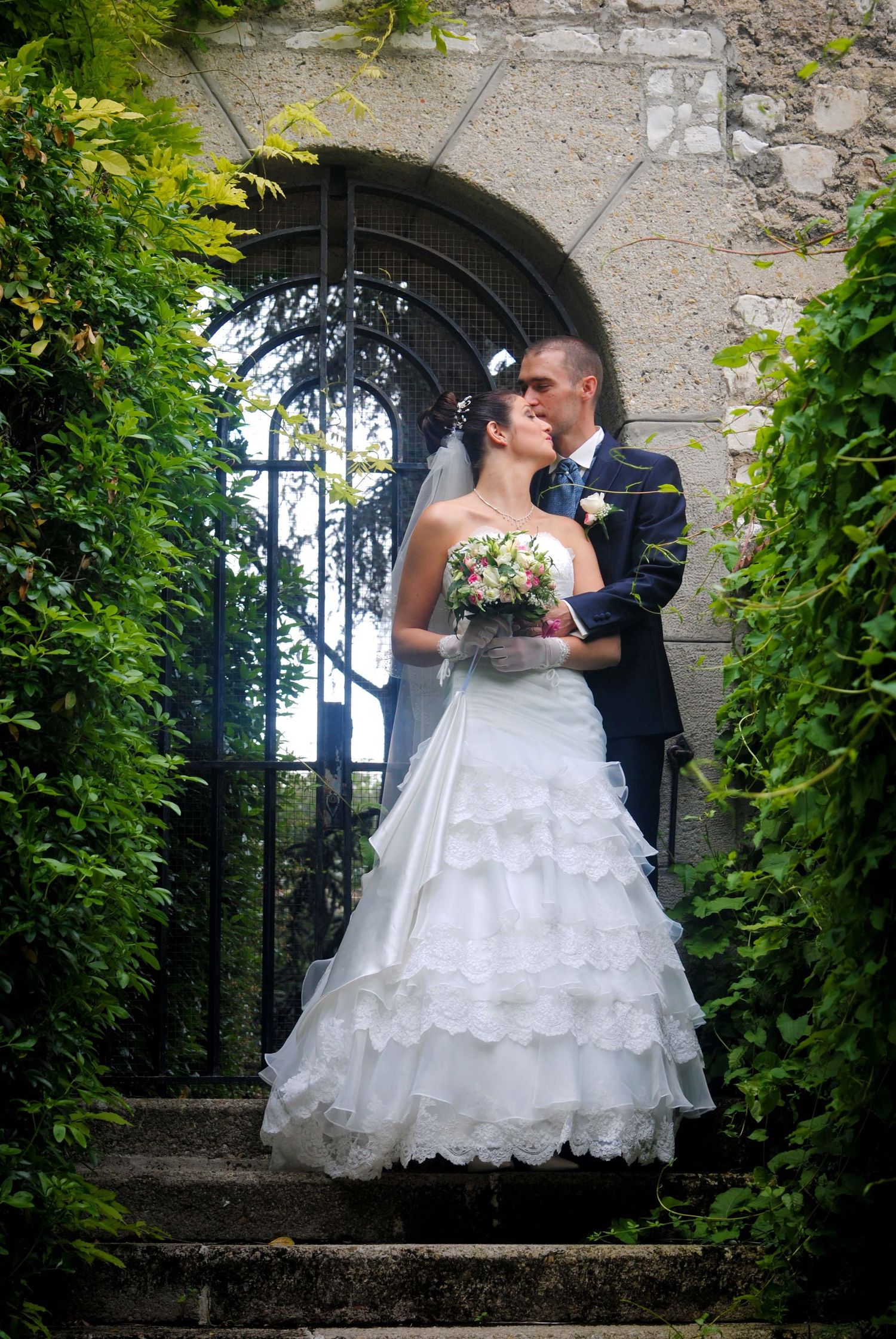 A bride in a ruffled white gown and groom in navy suit kissing at an arched garden gate.