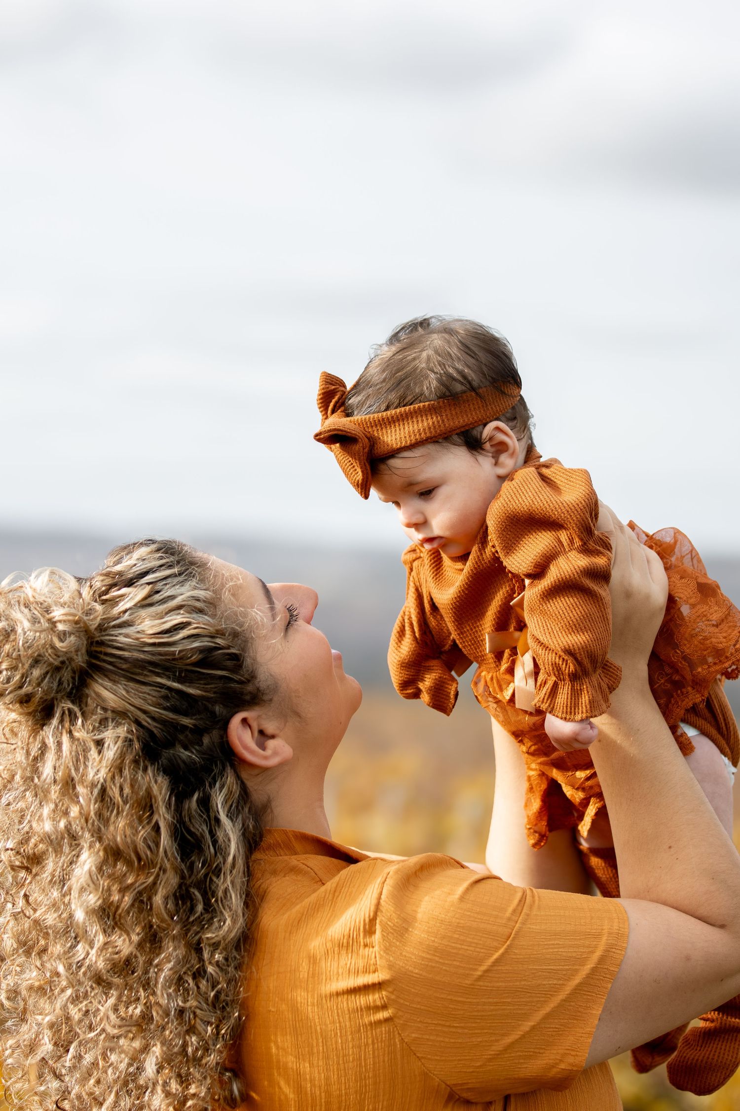 Curly-haired mother in orange dress lifting her baby girl in a matching rust outfit outdoors.