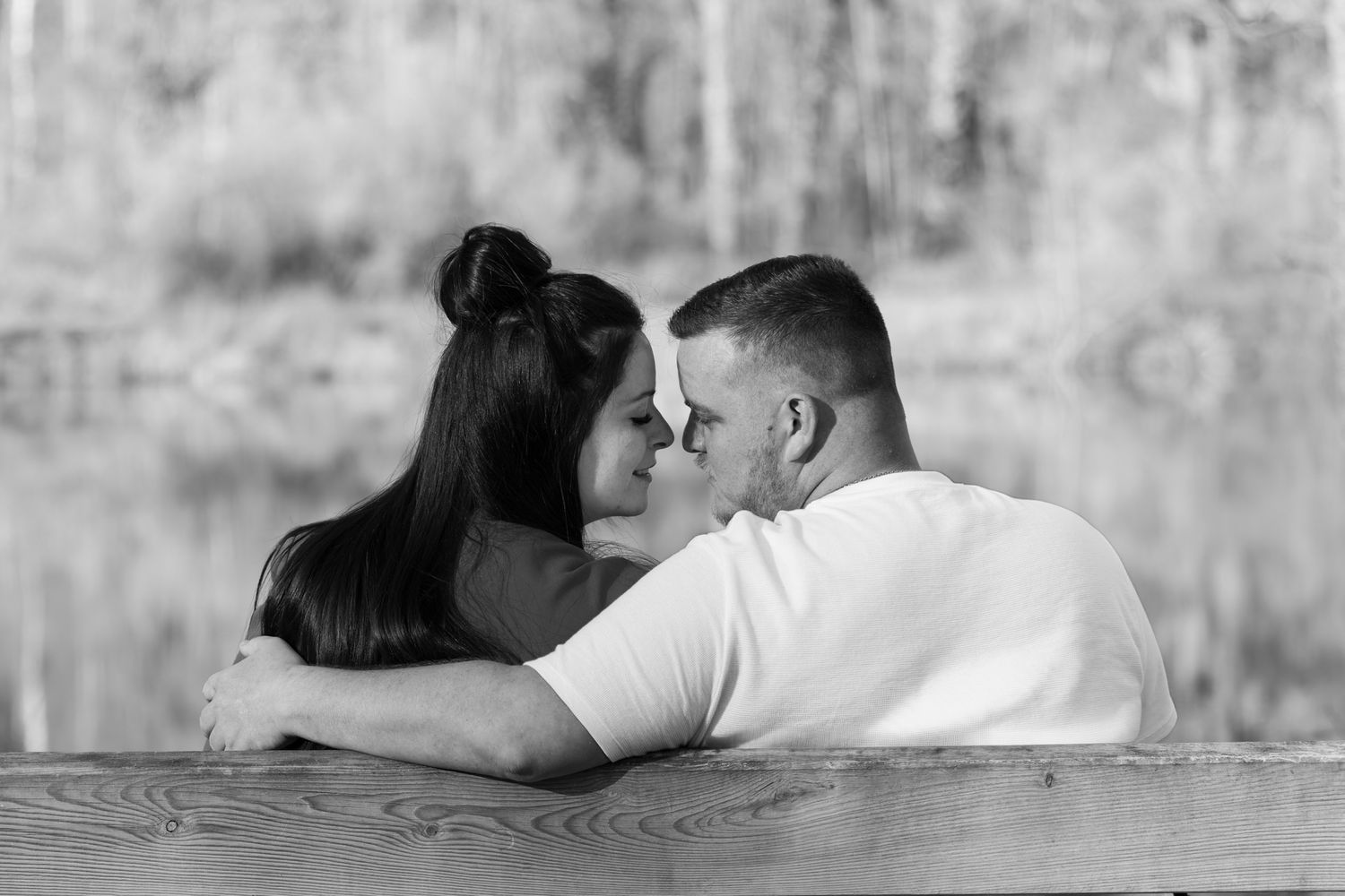 Black and white photo of a romantic couple leaning close together on a wooden dock near a serene lake.