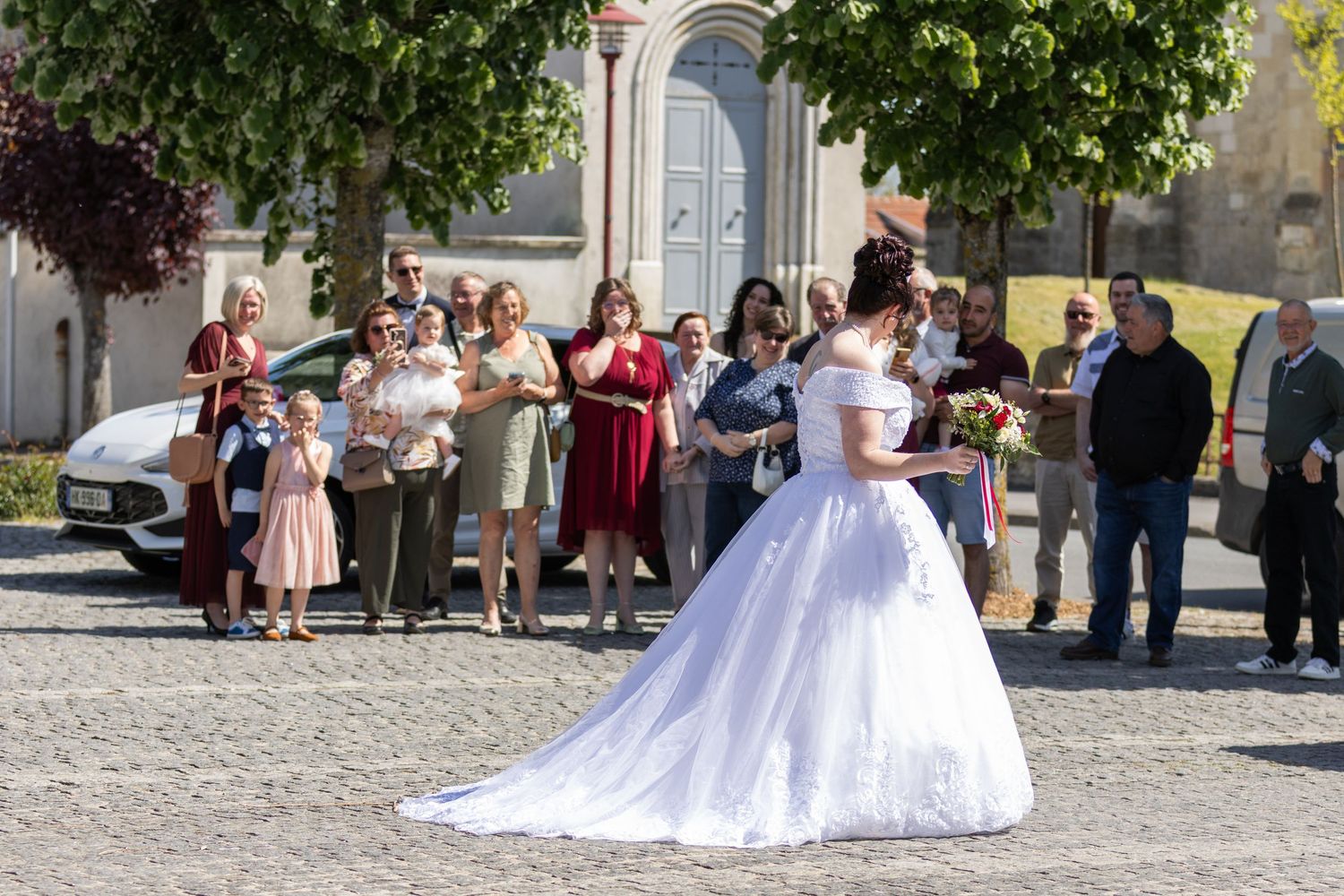Bride in white gown tosses bouquet to guests gathered outside a church on a sunny wedding day.