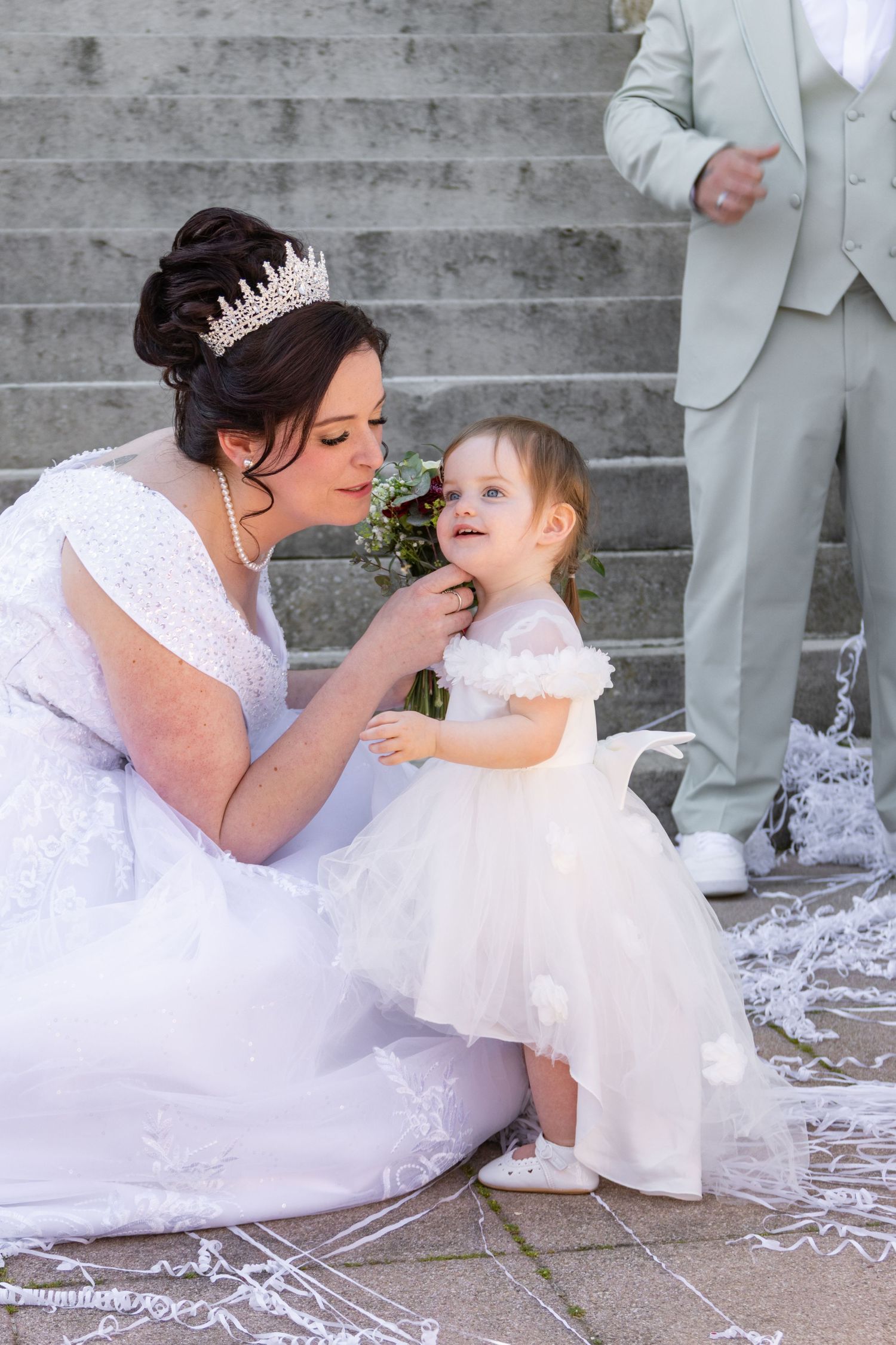 Bride in white gown and tiara kneels on steps with toddler flower girl in white dress during outdoor wedding ceremony.
