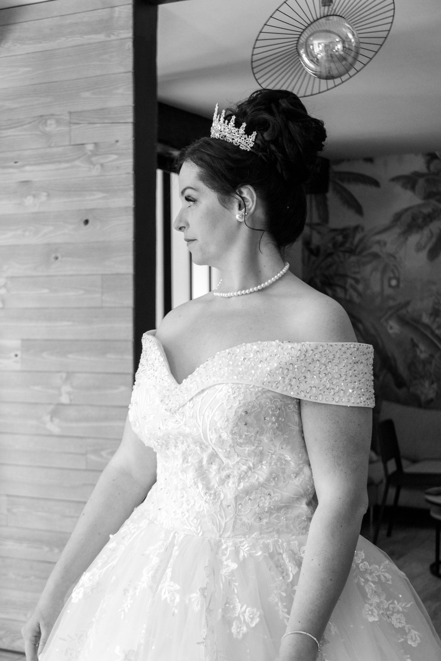 Black and white photo of a bride in an off-shoulder lace gown, pearl necklace, and tiara, gazing elegantly to the side.