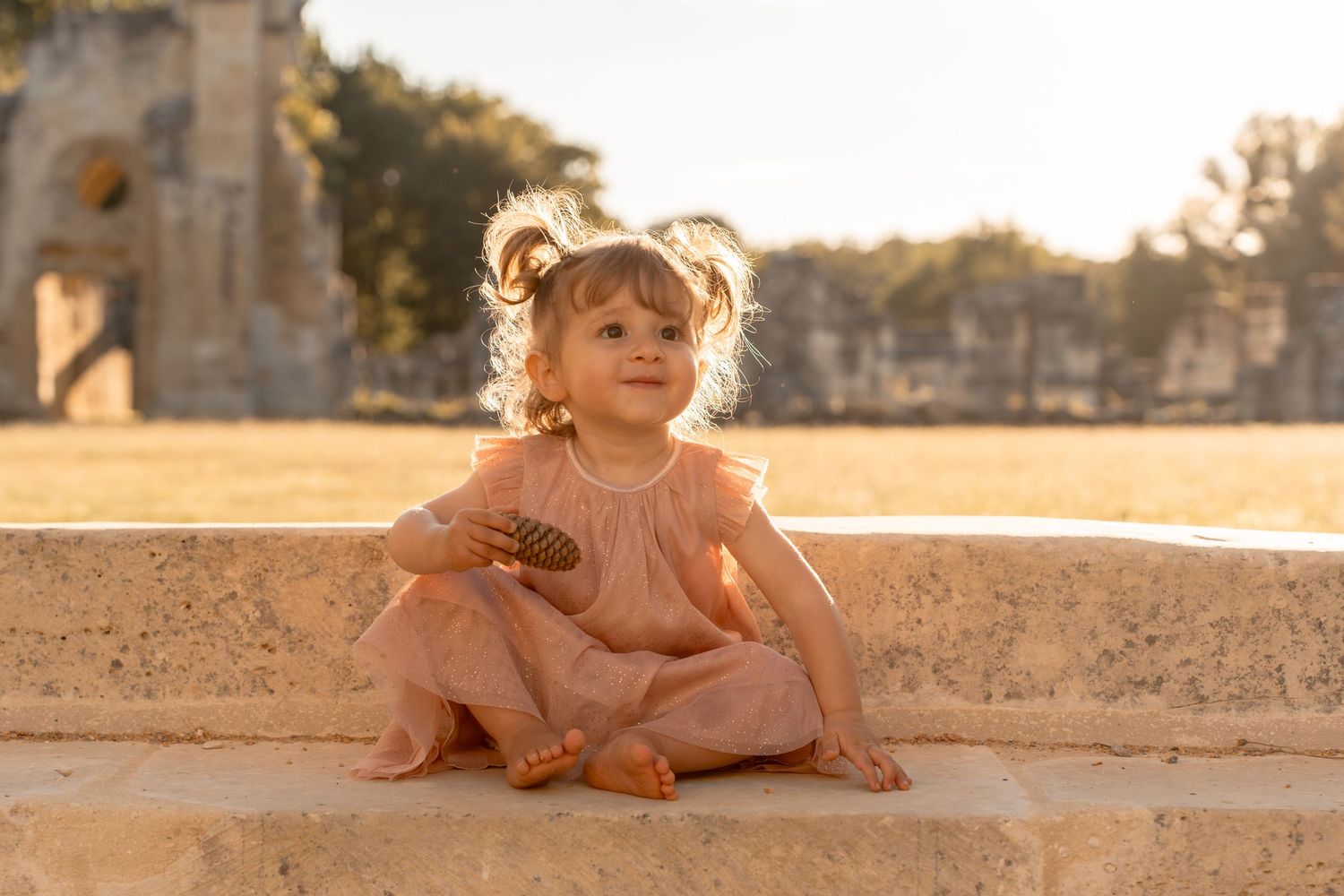 Adorable toddler girl in pink romper sitting cross-legged on stone steps holding a pinecone near ancient ruins.