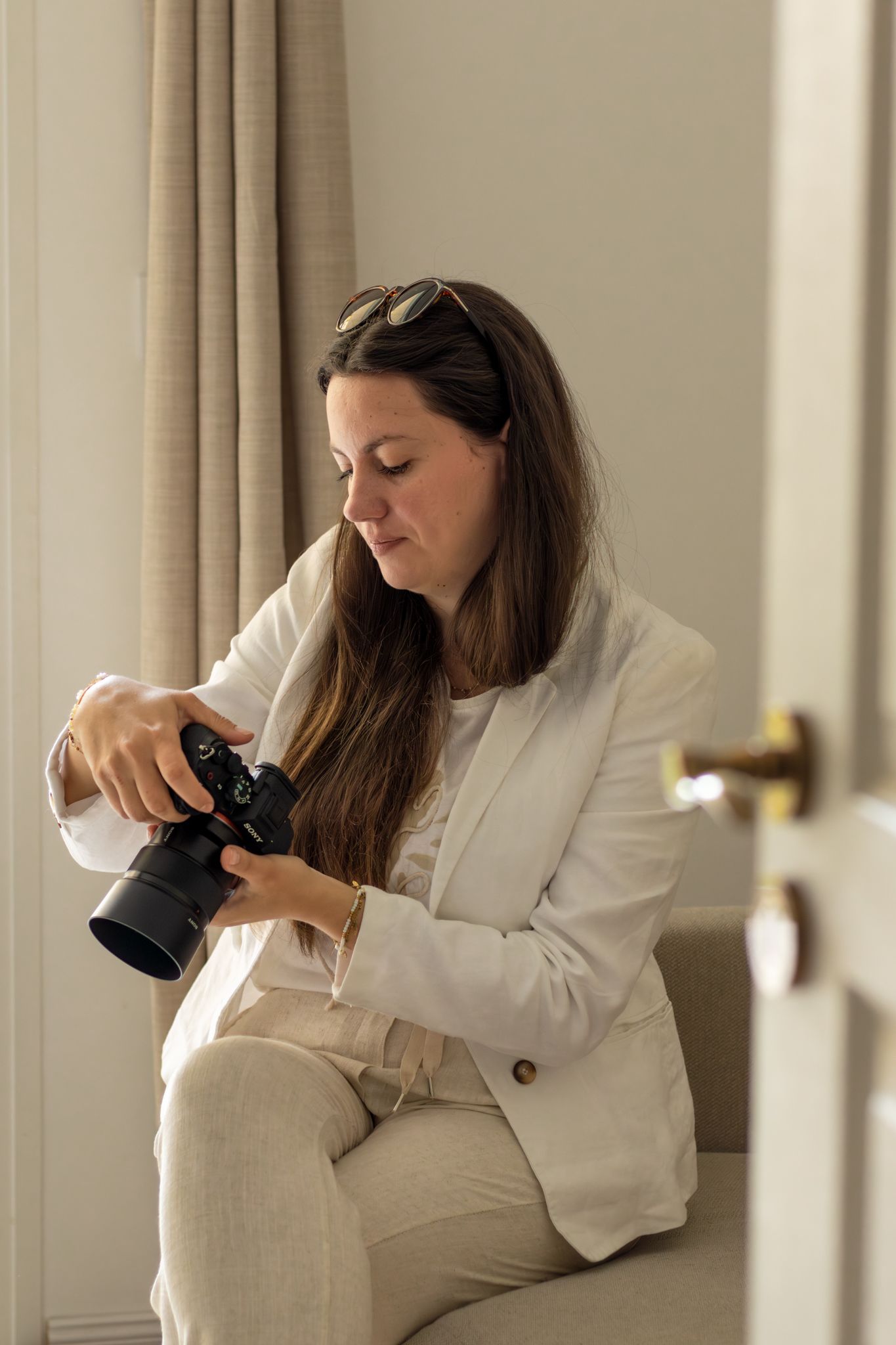 Woman in white blazer reviewing photos on a professional camera while seated indoors near a curtained window.