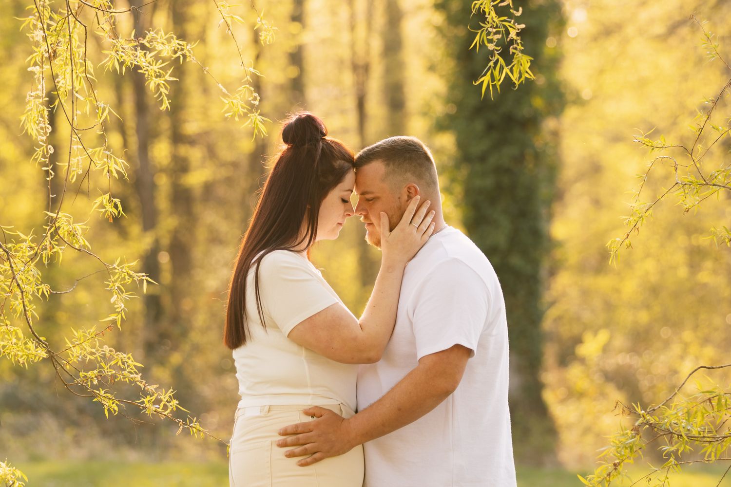 Pregnant couple in white embracing and touching foreheads in a sunny autumn park with golden foliage.