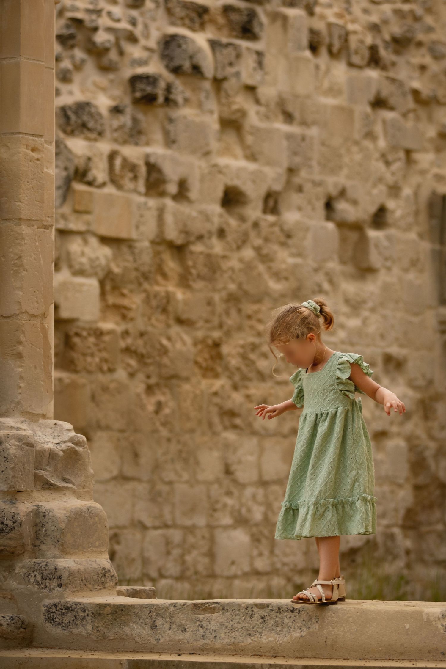 Young girl in green dress balancing on stone ledge beside ancient limestone wall.