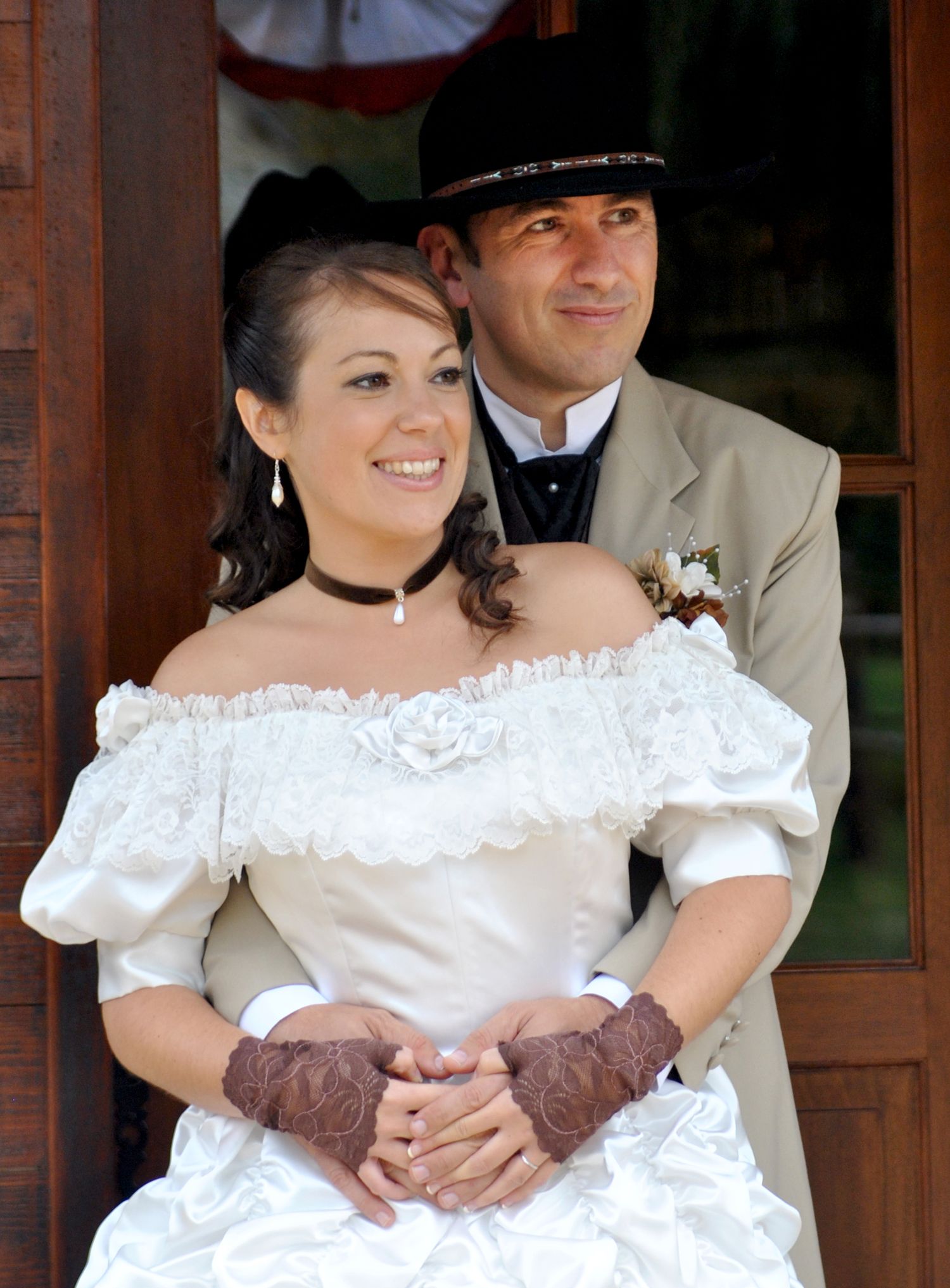 Western bride in white off-shoulder gown with brown lace gloves stands with groom in tan suit and cowboy hat.