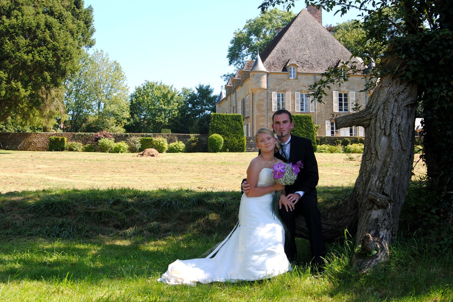 Bride and groom pose together by a tree in front of a charming French countryside manor on their wedding day.