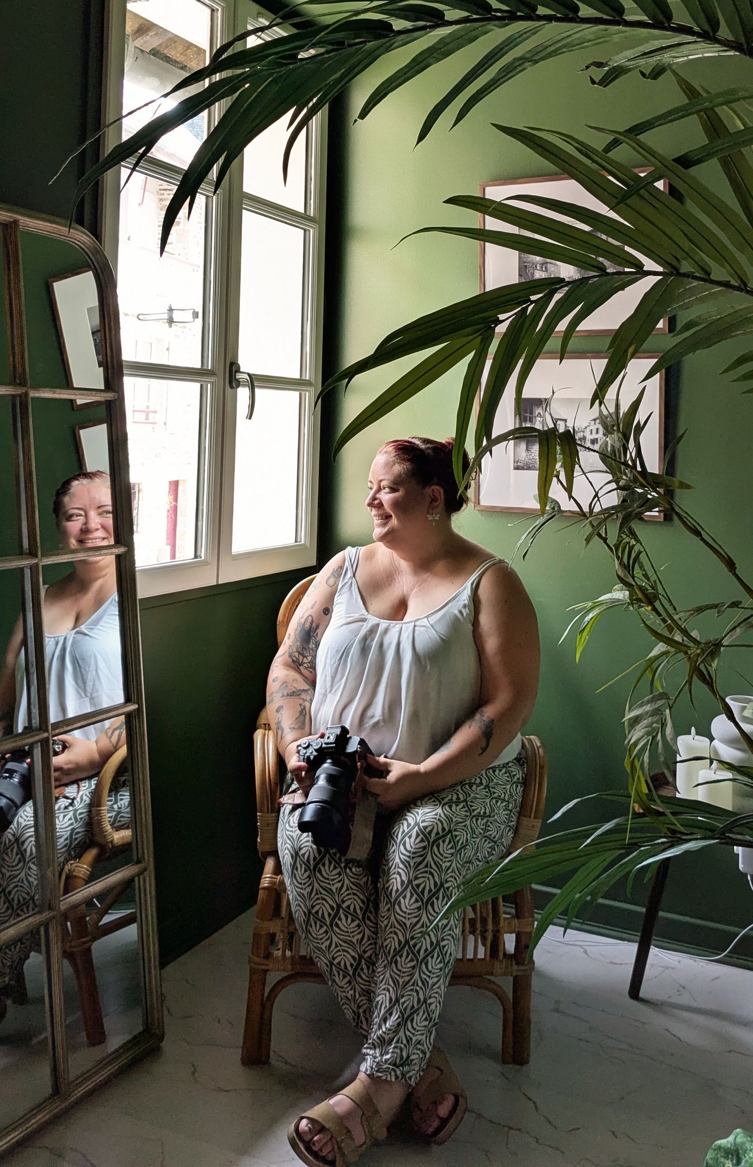 Woman with tattoos sits in rattan chair holding camera, reflected in mirror, surrounded by tropical plants on green wall.