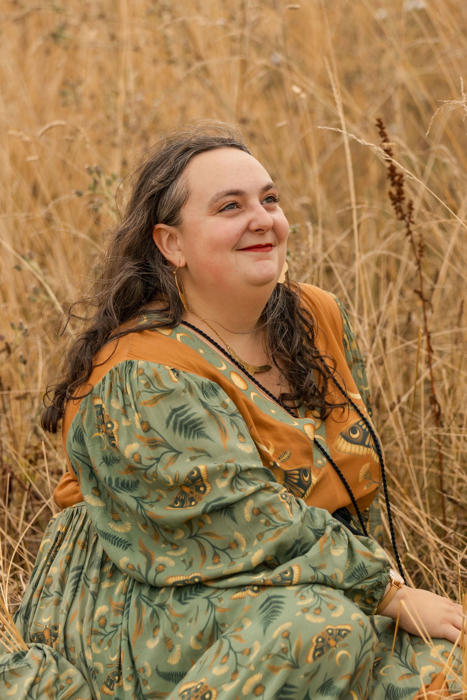 Smiling woman in green floral dress sitting in tall golden grass field.