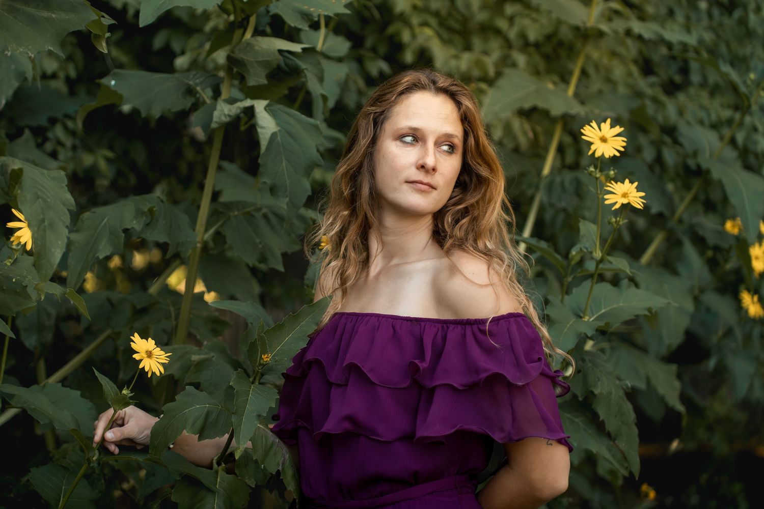 Woman in purple off-shoulder dress surrounded by green foliage and yellow flowers outdoors.